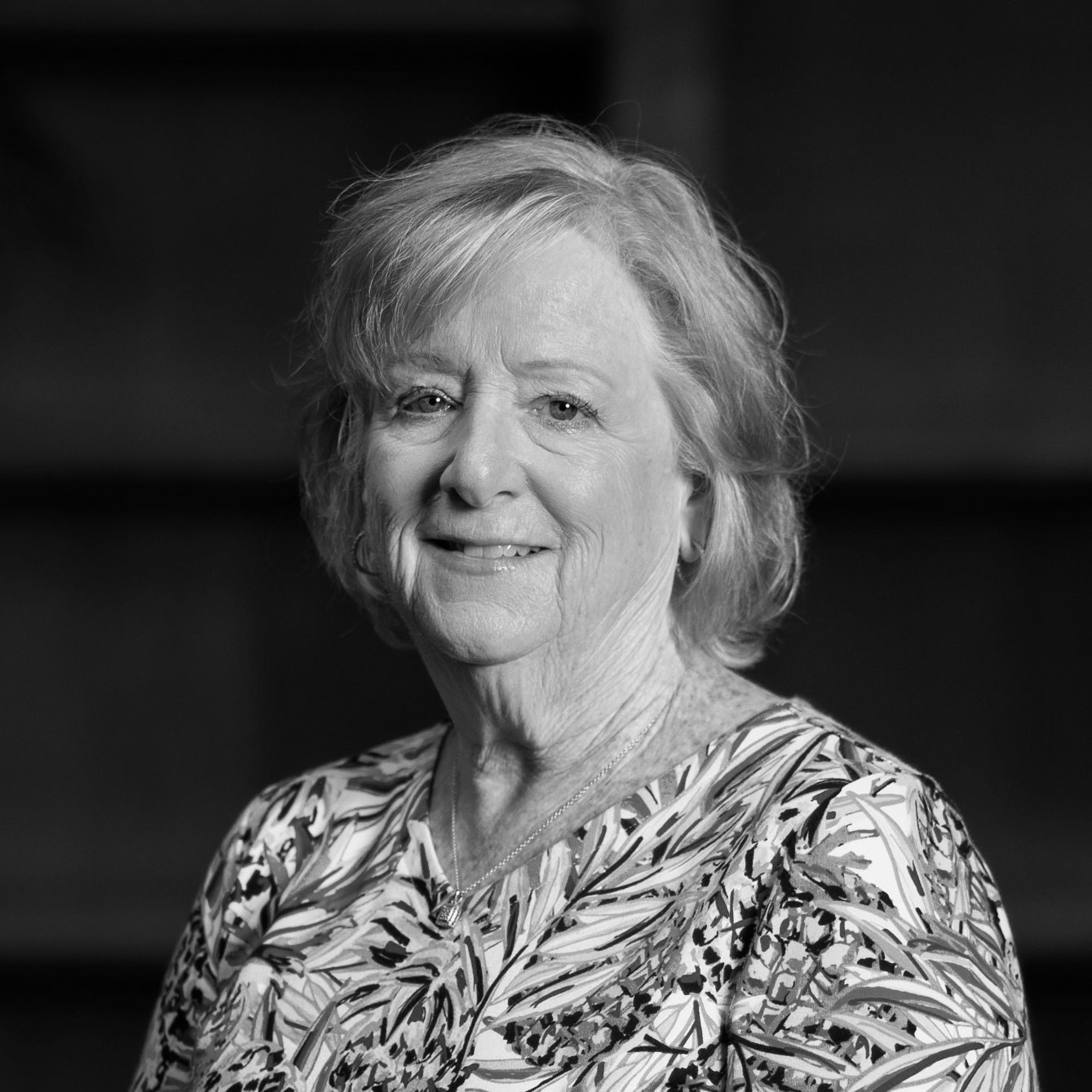 Black and white portrait of an elderly woman with short curly hair, wearing a patterned blouse and a necklace.