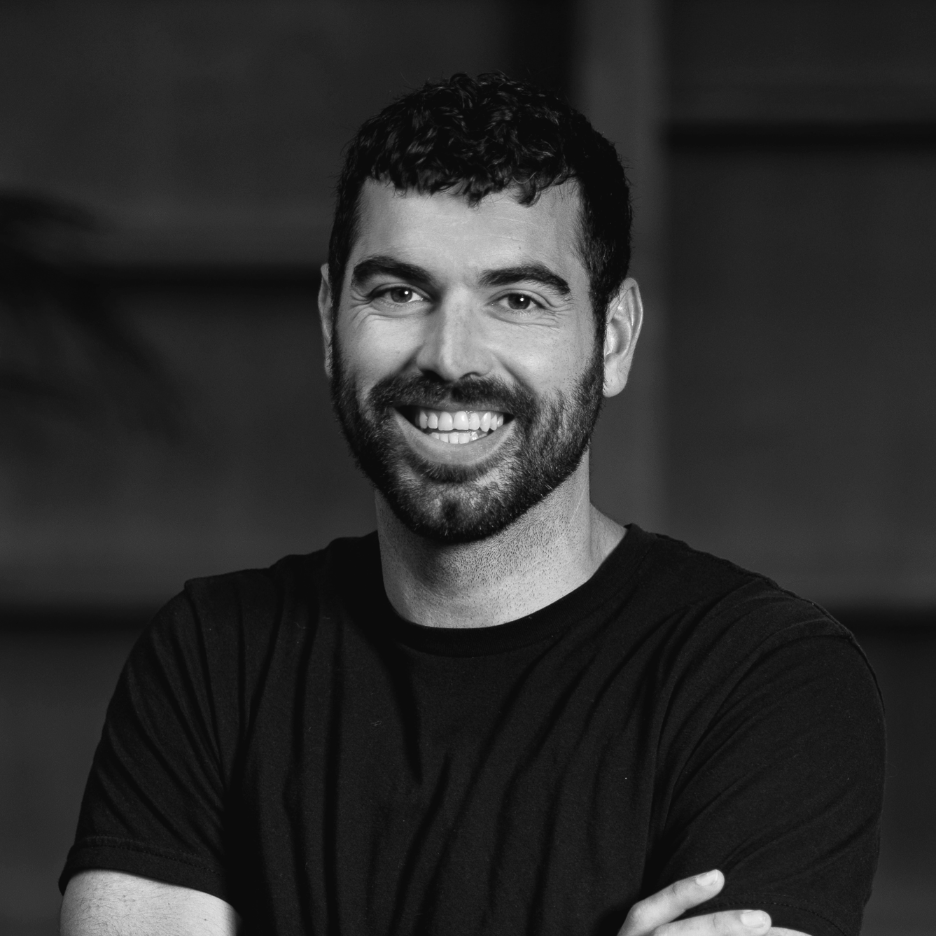Black and white portrait of a smiling man with dark hair and beard, wearing a black t-shirt, with arms crossed.