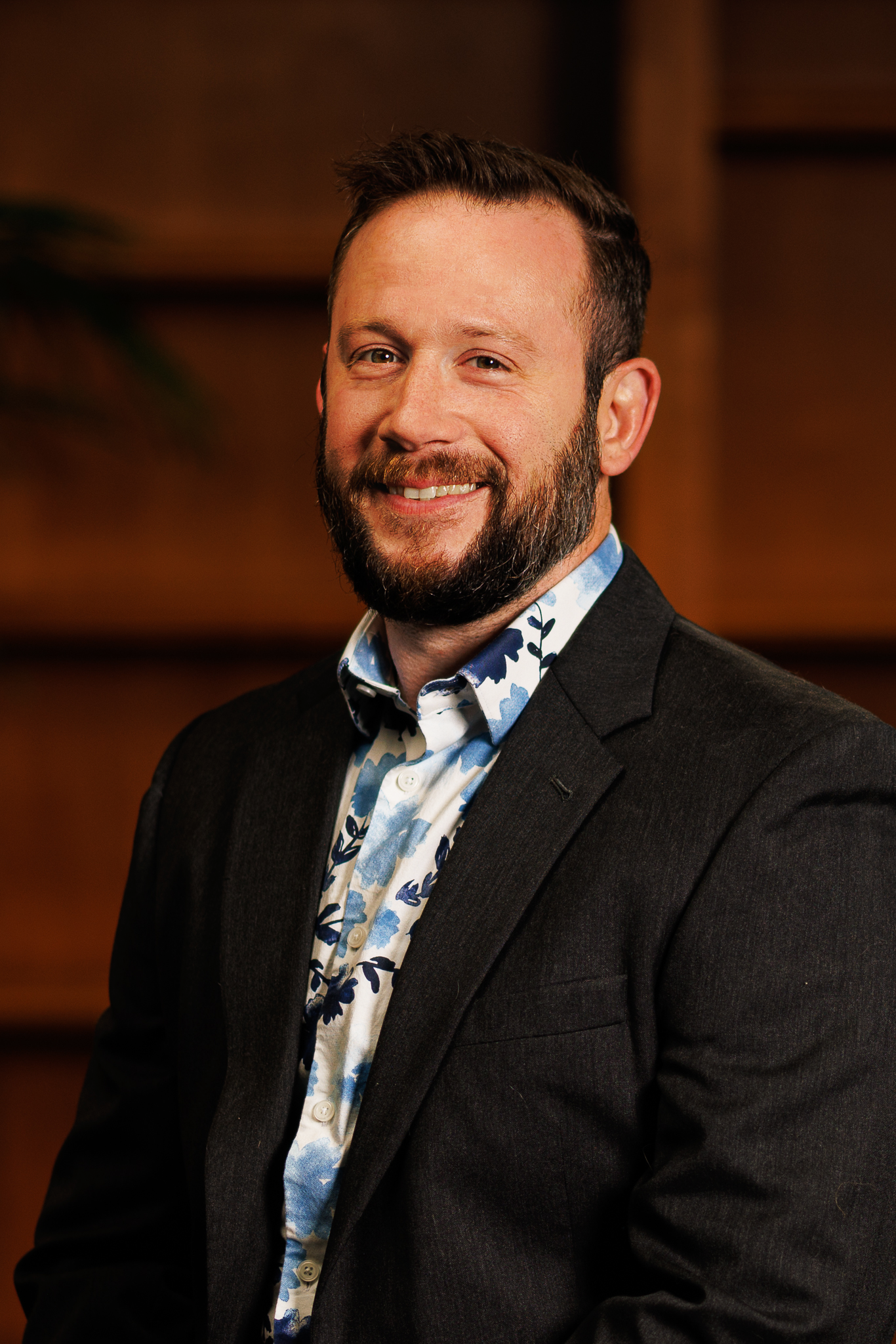 A smiling man with a beard in a dark blazer and a blue and white patterned shirt against a warm, blurred background.