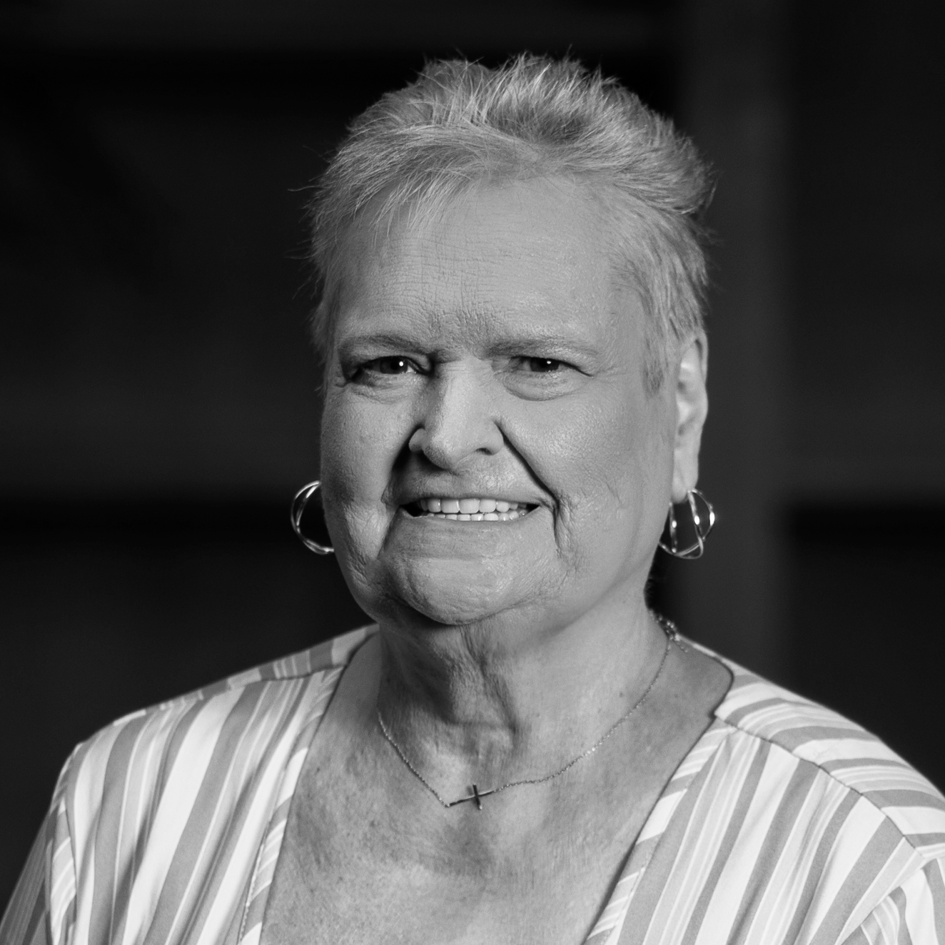 Black and white portrait of an older woman smiling, wearing earrings, a striped top, and a cross necklace.