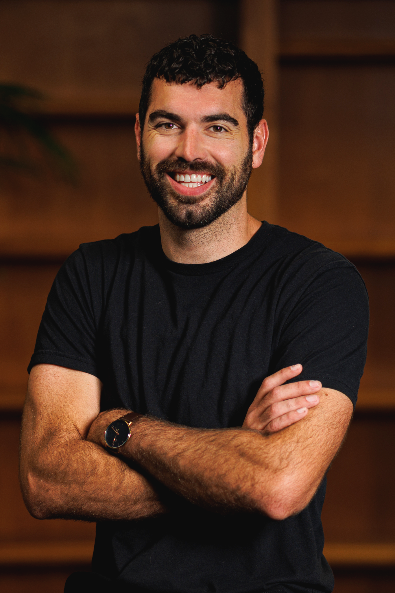 A smiling man with dark hair and a beard, wearing a black t-shirt and a watch, standing with arms crossed against a wooden background.