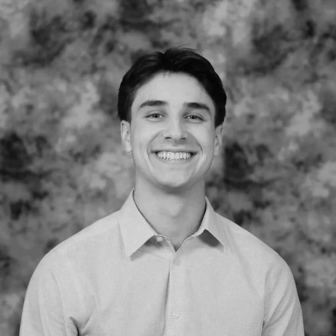 Black and white portrait of a young man with dark hair, smiling, wearing a collared shirt, with a textured background.