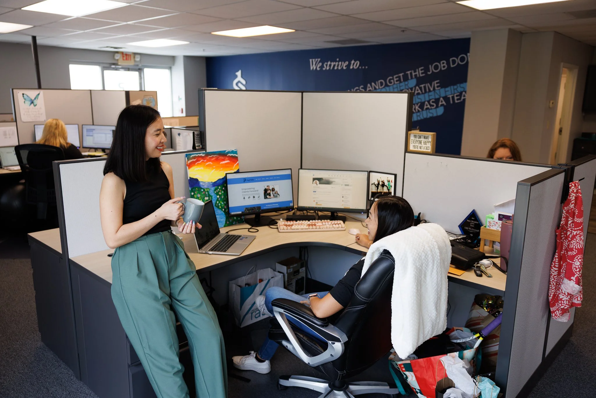 Two women talking in an office cubicle with computers and office supplies.