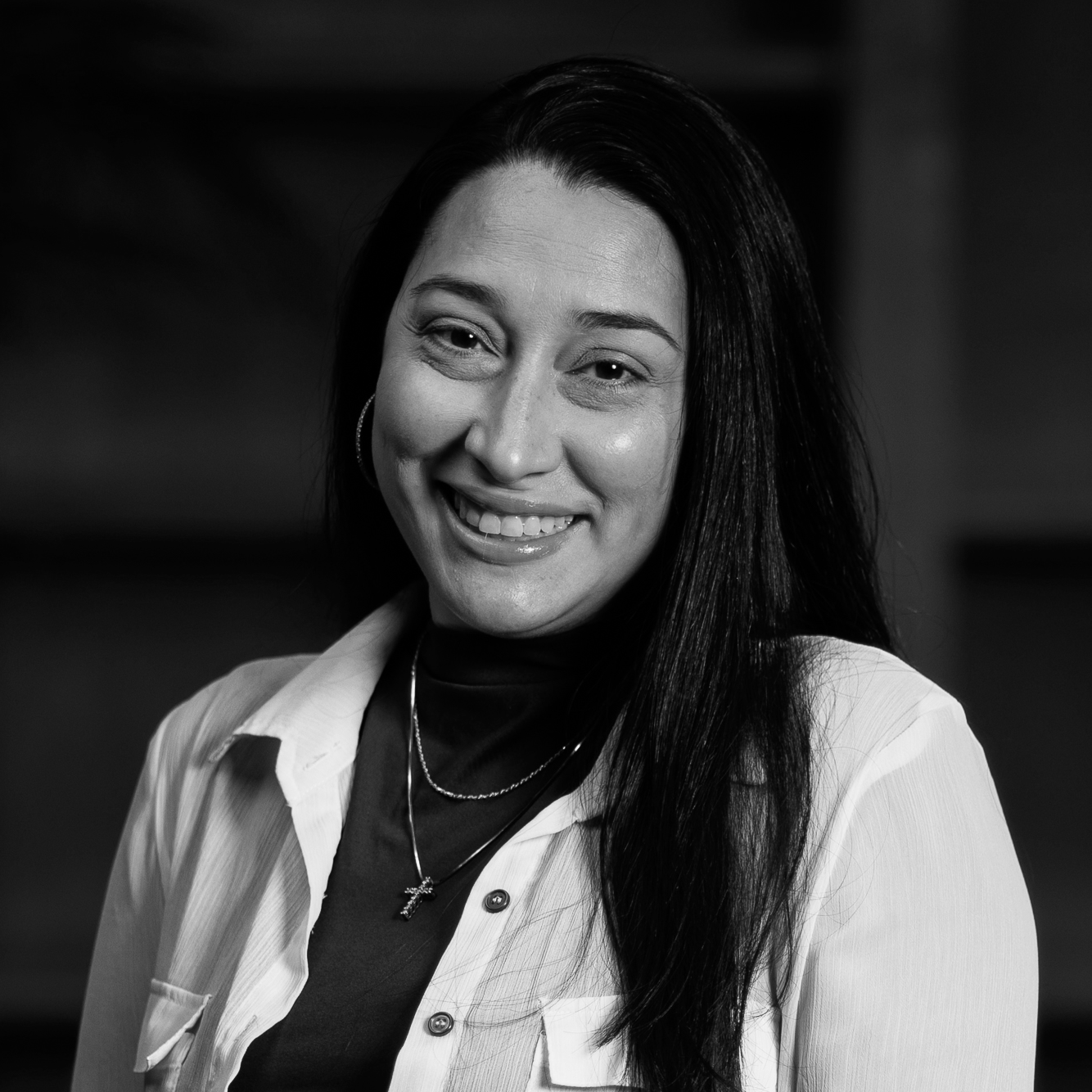 A smiling woman with long dark hair, wearing hoop earrings, layered necklaces, a dark top, and a light-colored shirt.