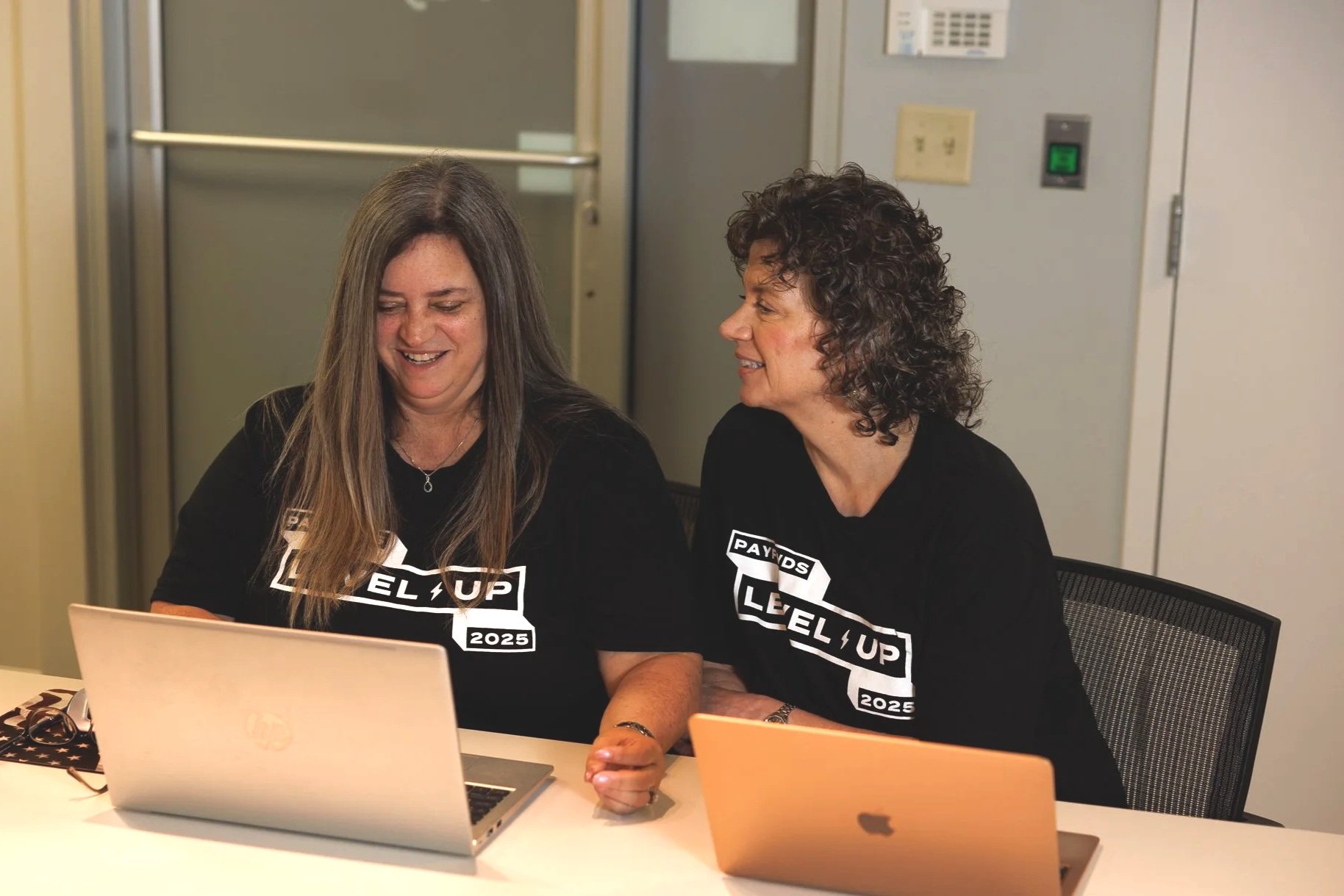 Two women sitting at a table with laptops, smiling and looking at one of the screens, wearing matching black t-shirts with a 'Level Up 2025' logo.