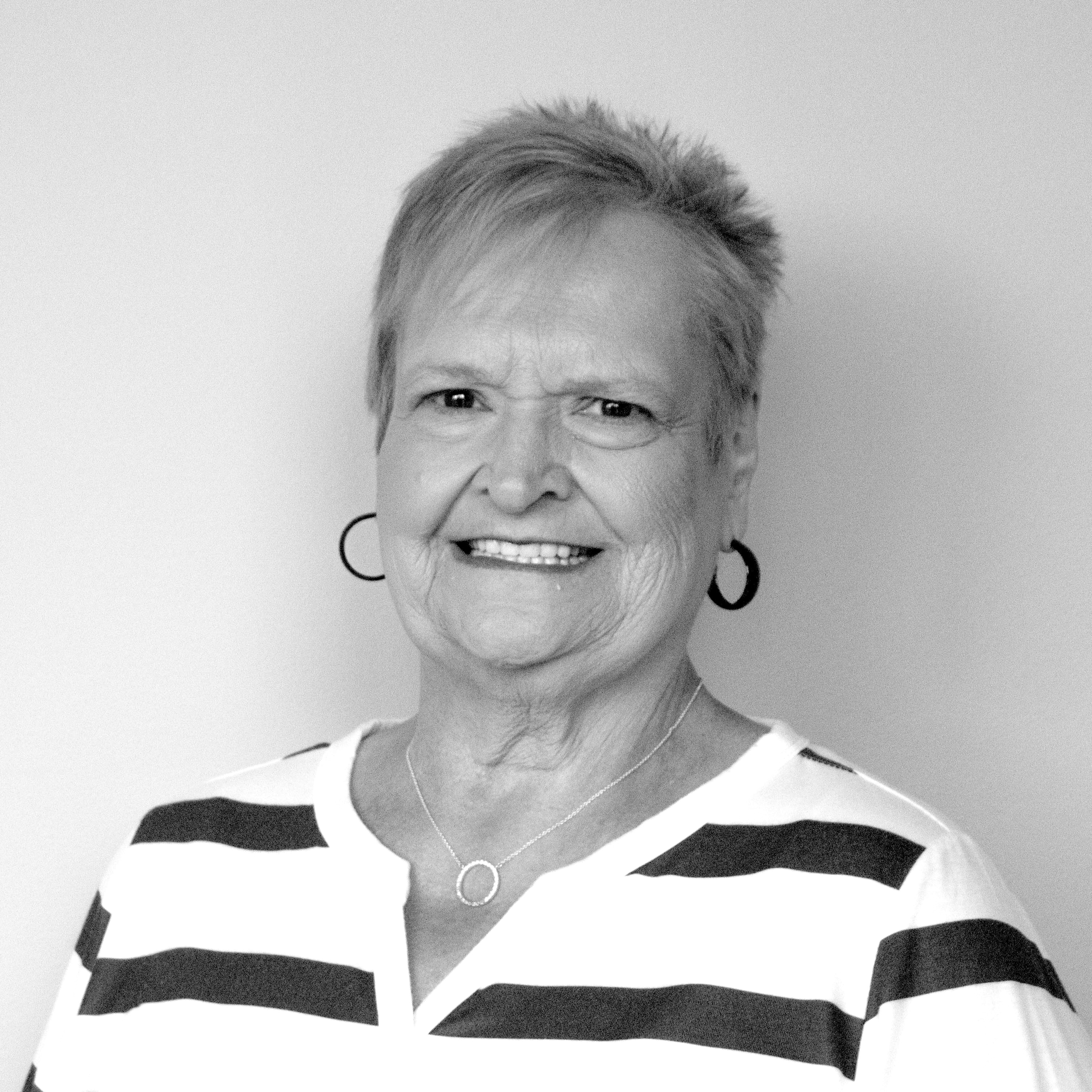 Black and white portrait of an older woman smiling, wearing earrings, a striped top, and a cross necklace.