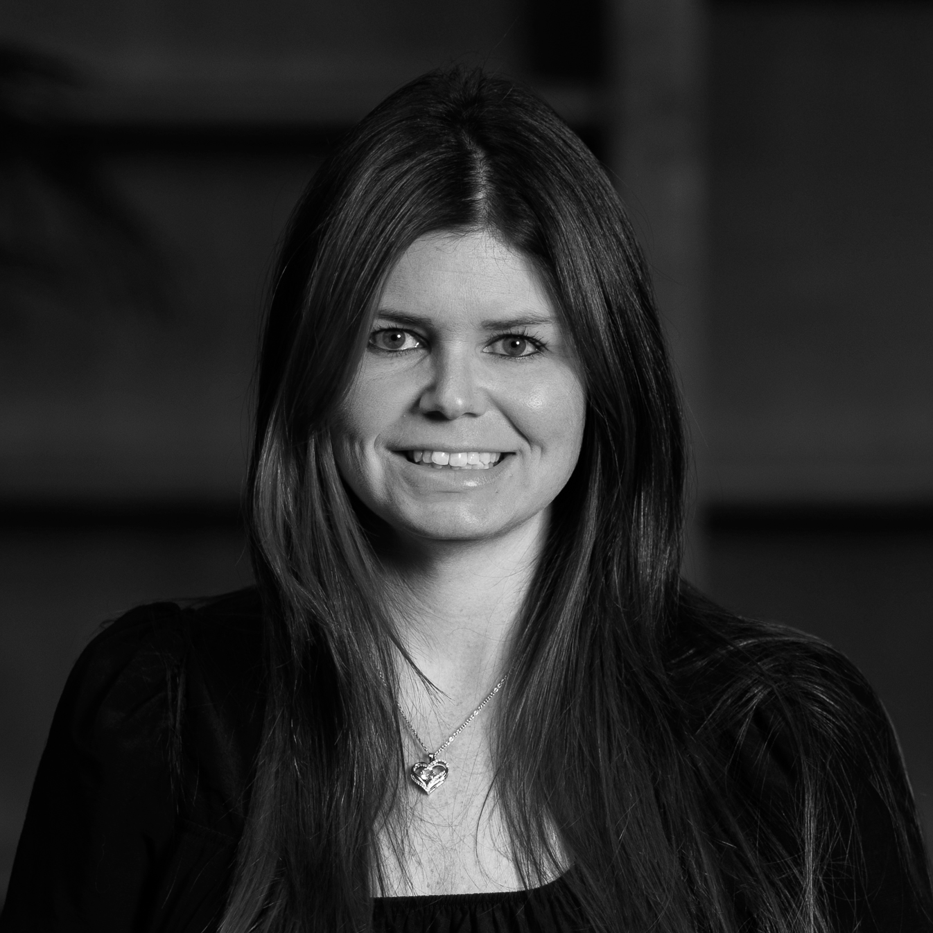 Black and white portrait of a smiling woman with long dark hair, wearing a necklace with a heart-shaped pendant.