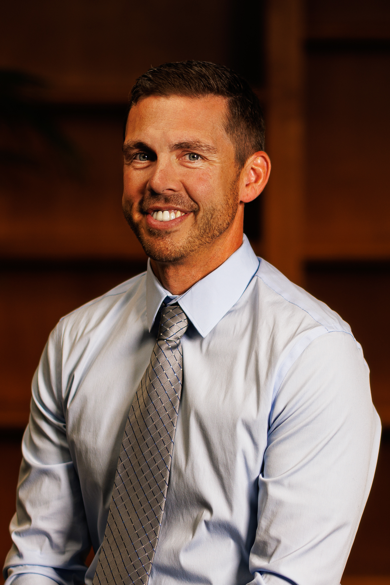 A smiling man with dark hair, wearing a light blue dress shirt and a plaid tie, in a professional setting with a wooden background.