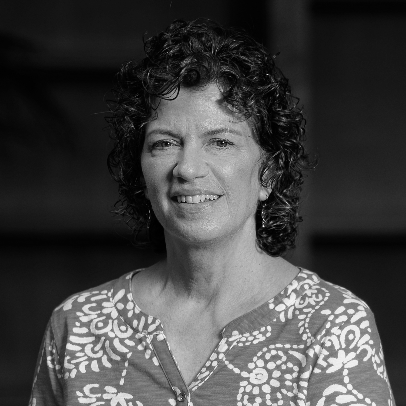 Black and white portrait of a woman with curly short hair, wearing earrings and a patterned blouse, smiling slightly.