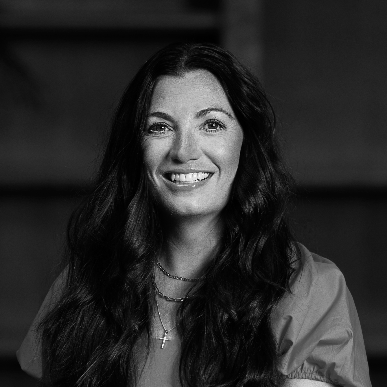 A smiling woman with long, wavy dark hair, wearing a light-colored top and layered necklaces, in a black and white portrait.