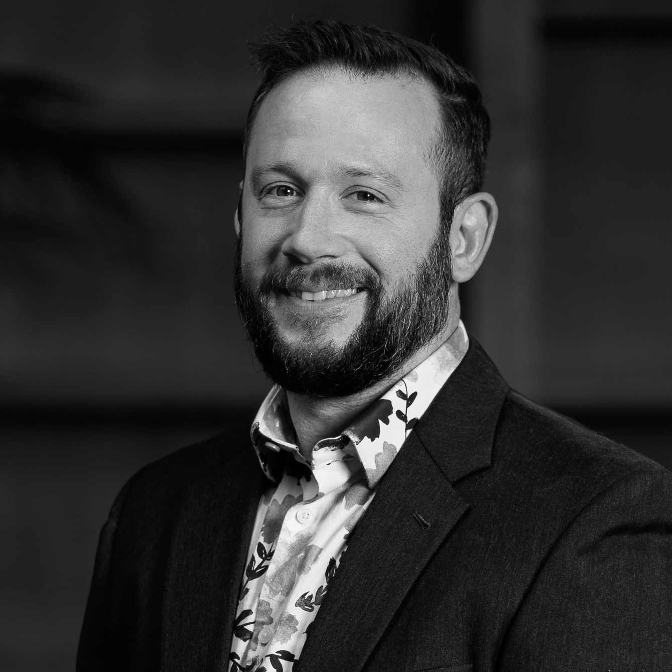 Black and white portrait of a smiling man with a beard, wearing a floral shirt and a blazer.