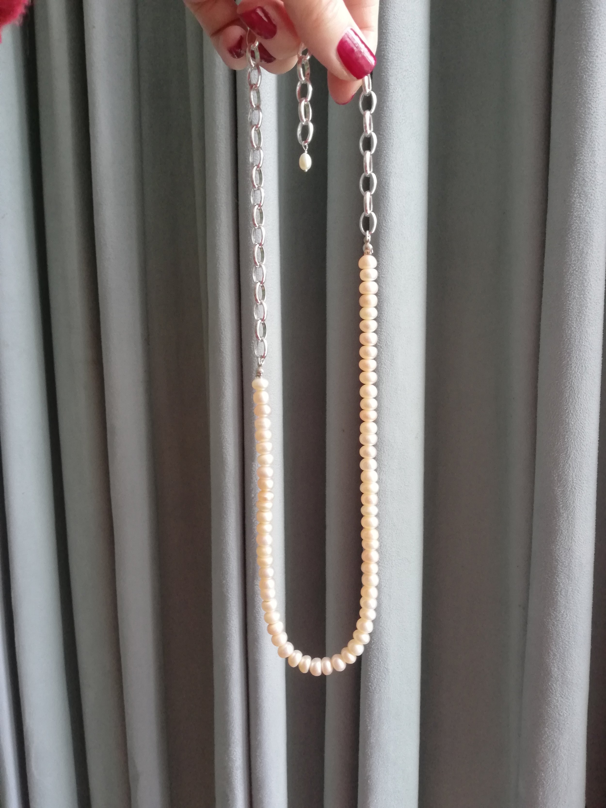 Hand holding a pearl necklace with a silver chain against a gray curtain backdrop.