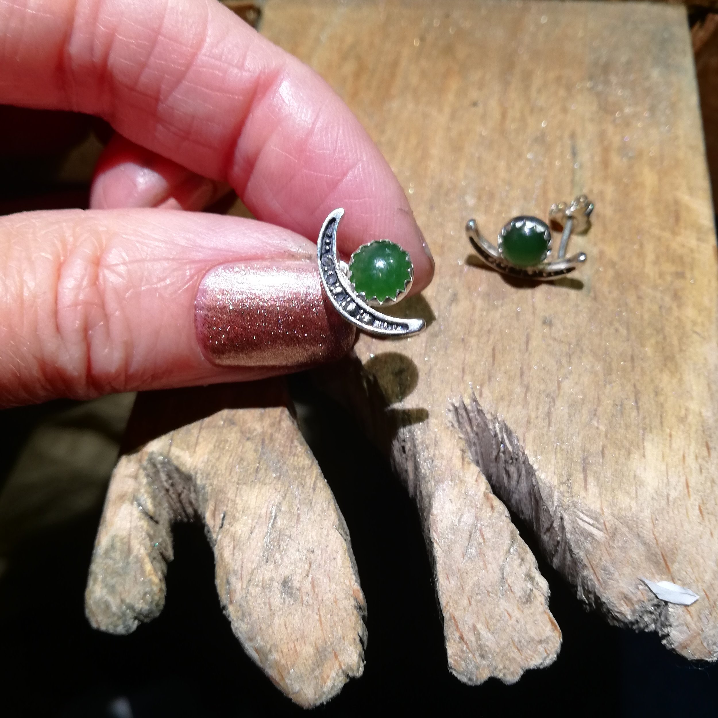 A person holding a silver earring with a green stone and crescent moon design, next to a matching earring on a wooden surface.