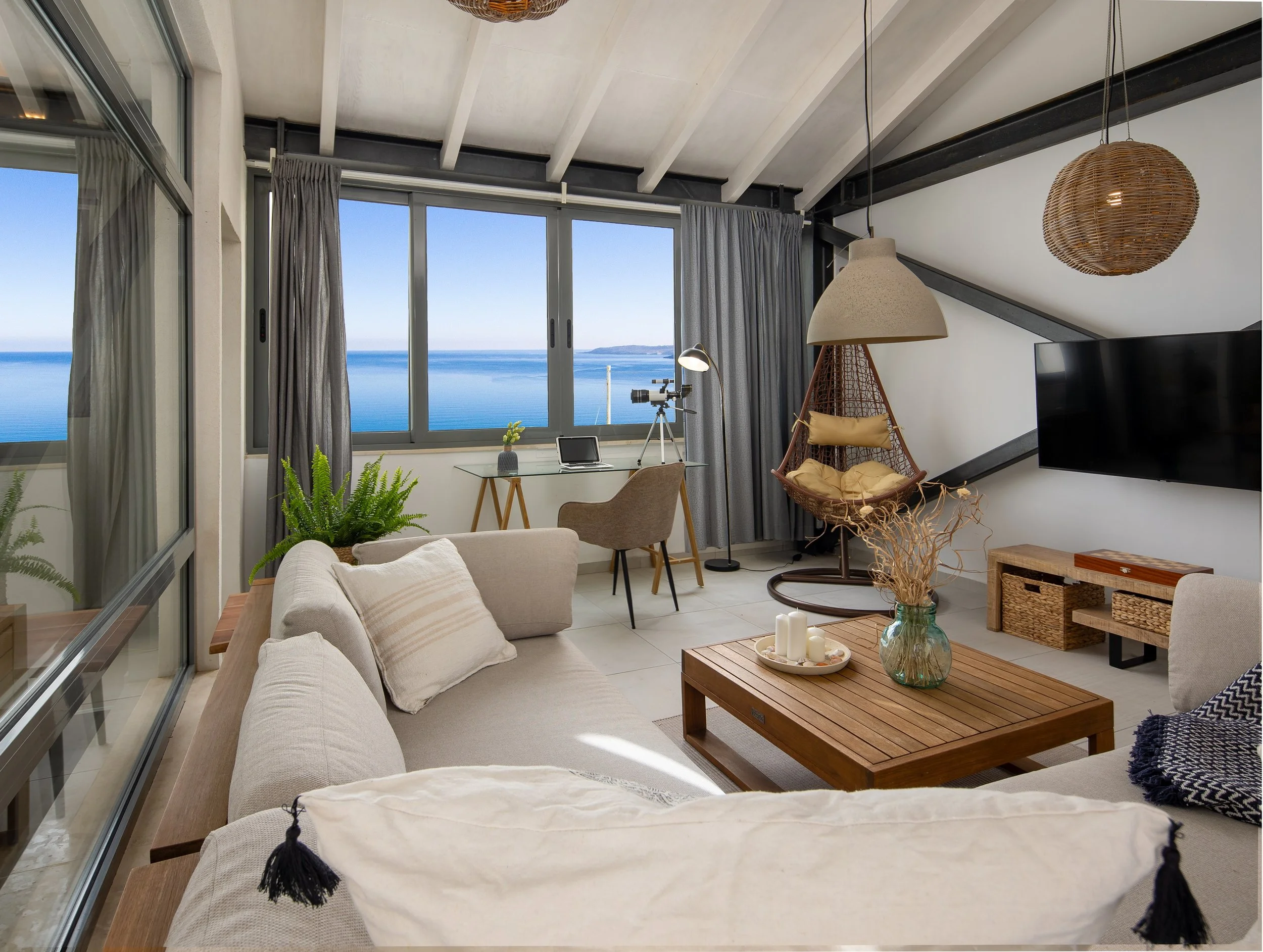 Bright living room with an amazing sea view, featuring a large sofa, TV, a stylish hanging chair, and a desk.