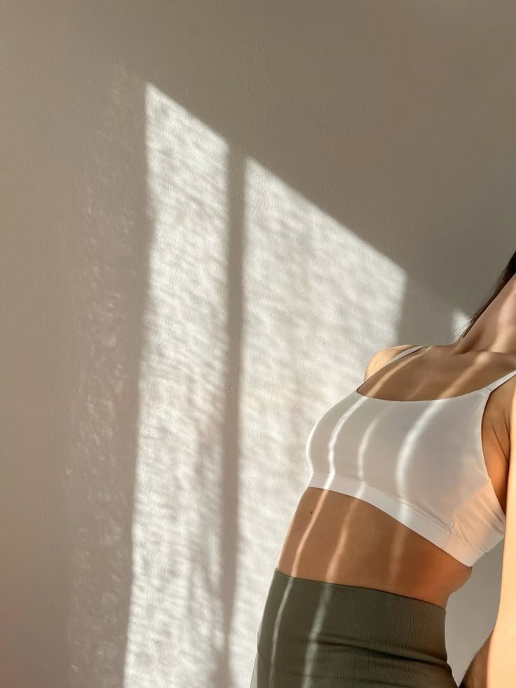 A woman standing near a wall with sunlight creating striped shadows on her body, wearing a white sports bra and dark high-waisted pants.