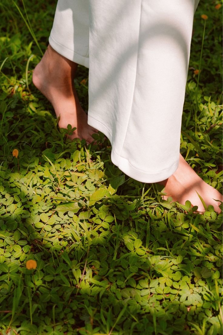 Person standing barefoot on green leafy ground, wearing white pants.