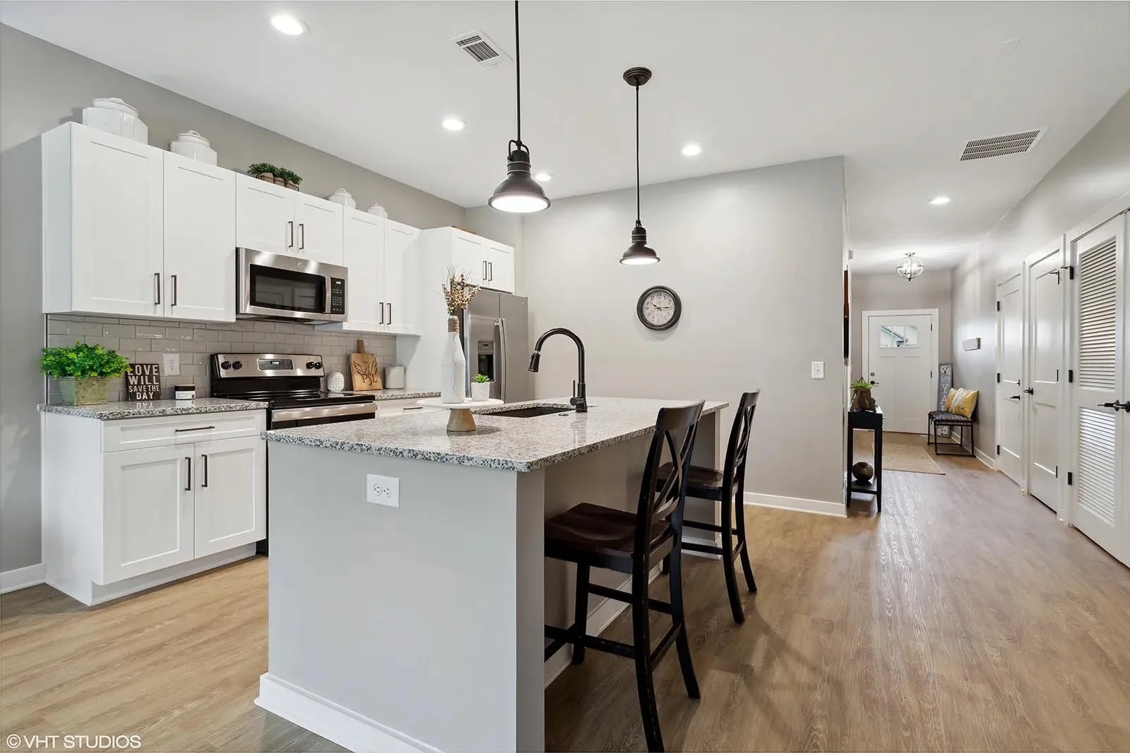 Modern kitchen with white cabinets, stainless steel appliances, granite countertops, an island with a black faucet, and wooden flooring. There are two black chairs at the island, and a hallway with a bench and decorative pillows in the background.