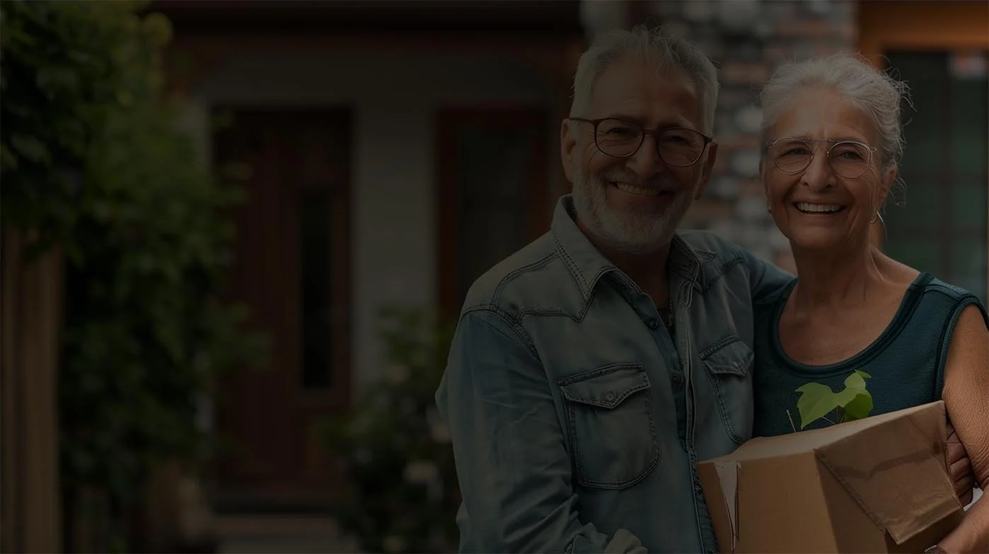 Smiling couple holding a cardboard box with a plant inside, standing outside a house with a wooden door. Happy to be downsizing!