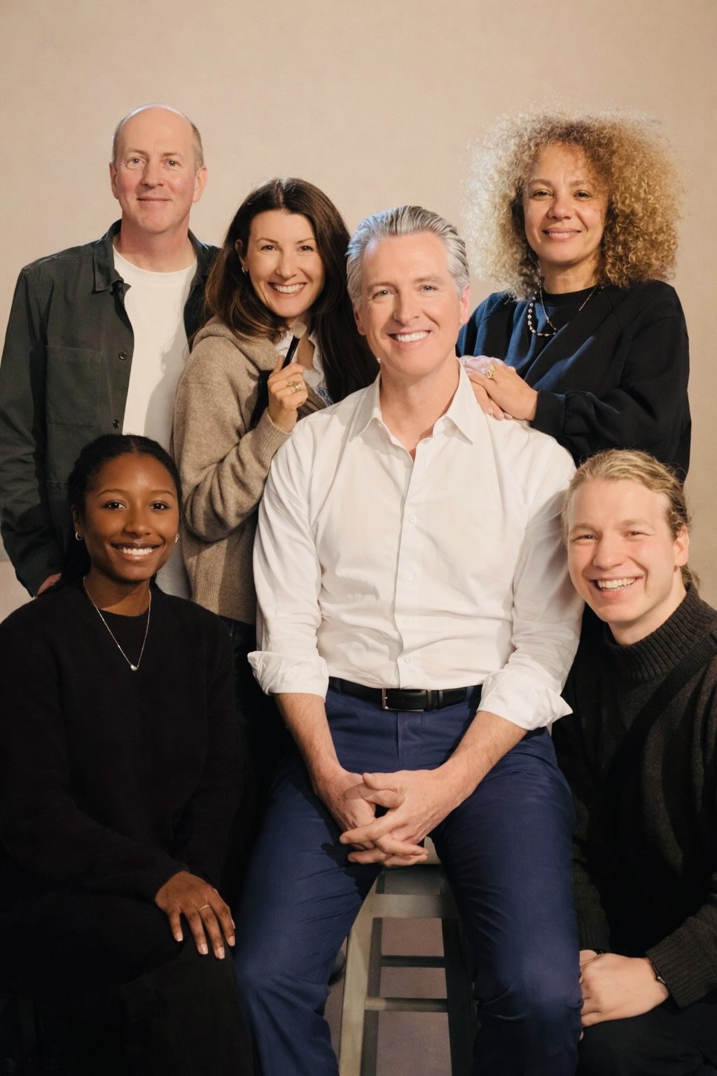 Group of seven diverse people smiling and posing for a photo indoors, some seated and others standing behind, dressed in casual and semi-formal clothes.