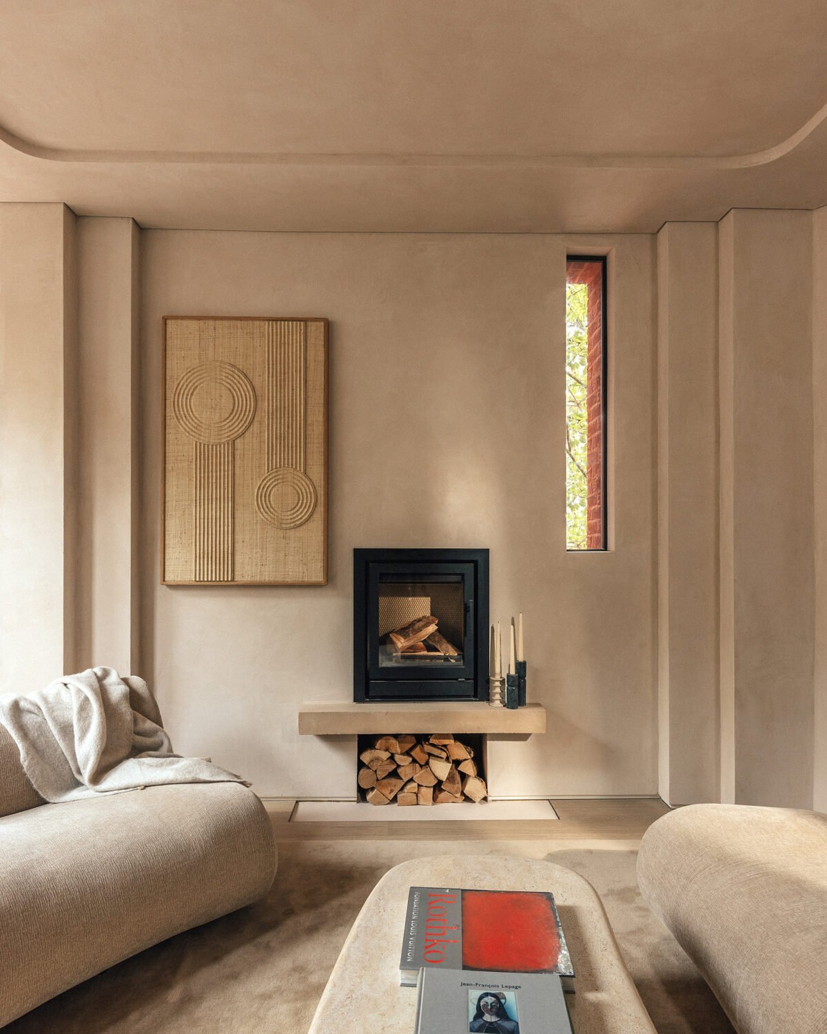 Living room with a beige sofa, abstract wall art, a black fireplace, a narrow window showing outdoor greenery, and a wood storage area underneath the fireplace, with candles on the mantle.