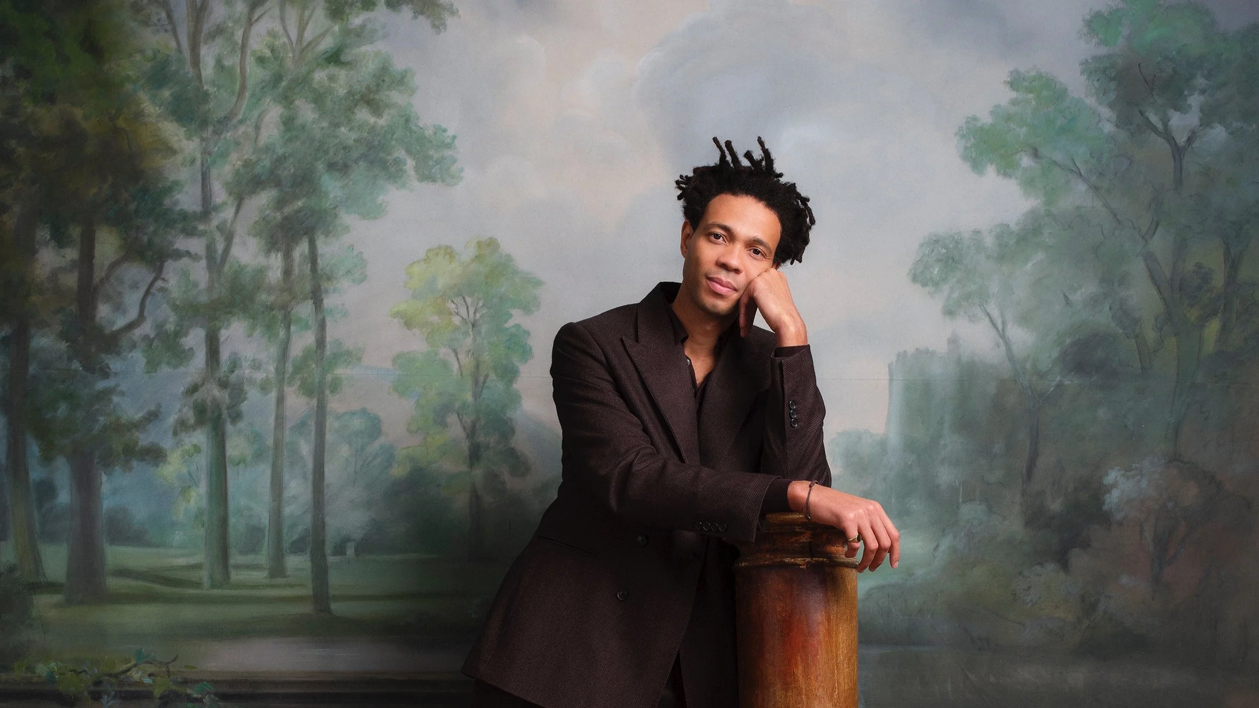 A man with dark curly hair in a brown suit poses with his head resting on his hand, standing in front of a painted forest background.