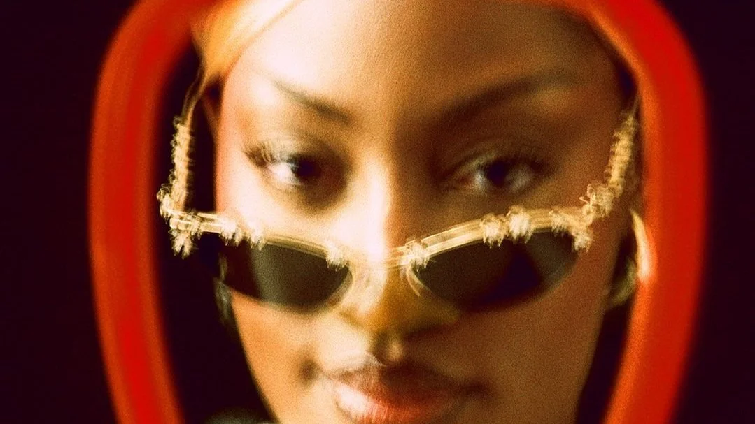 Close-up of a woman wearing sunglasses, viewed through a mirror with an orange frame, with makeup and platinum blonde hair.
