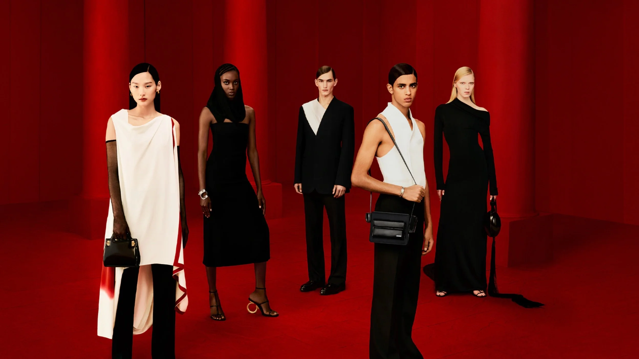 Group of five models standing against a red background, wearing high fashion black and white outfits, holding handbags and wearing heels.