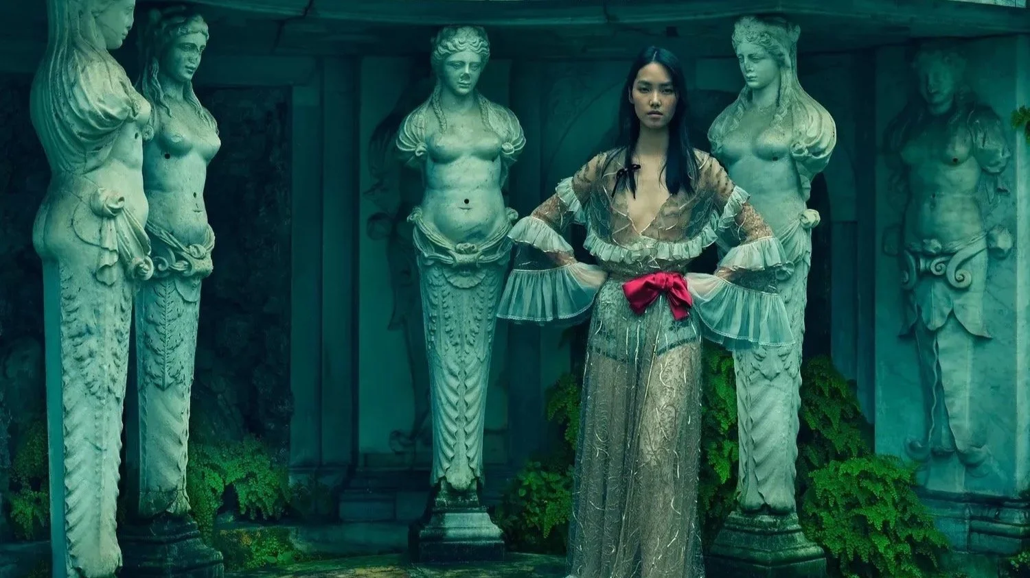 A woman standing among classical Greek statues with her hands on her hips, wearing a sheer dress with ruffled sleeves and a red ribbon belt.