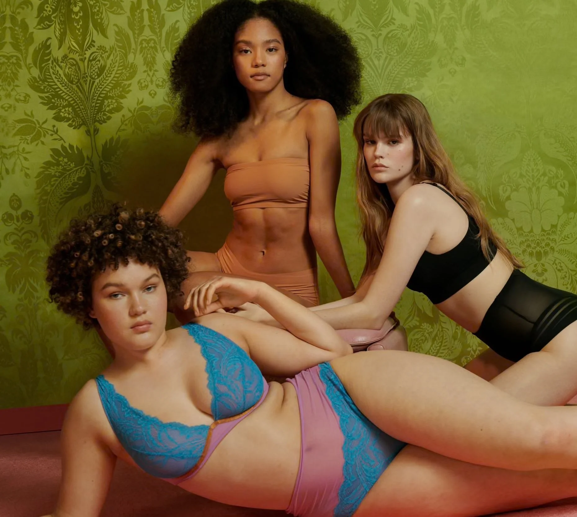 Four women with different hair colors and textures posing in lingerie in front of a green patterned wall.