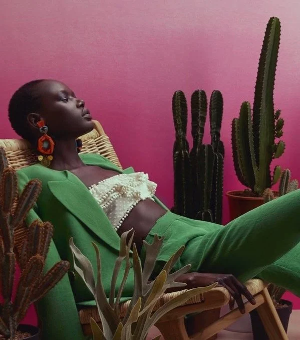 A woman with dark skin, wearing a bright green suit and large colorful earrings, reclining in a wicker chair with a pink wall and several tall cacti in pots behind her.