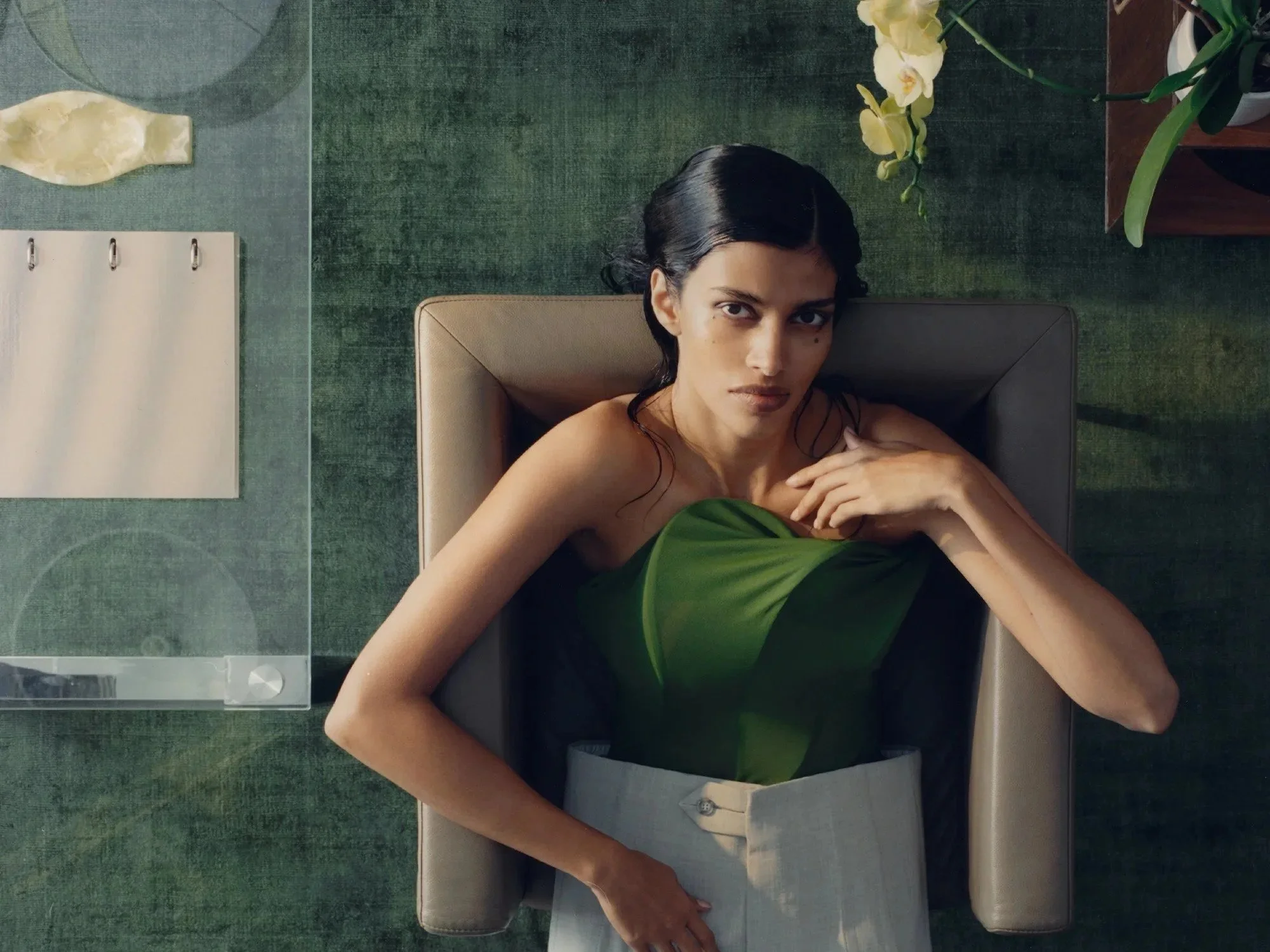 A woman with dark hair and skin tone lying on a bed, looking directly at the camera, with plants and a green textured wall in the background.