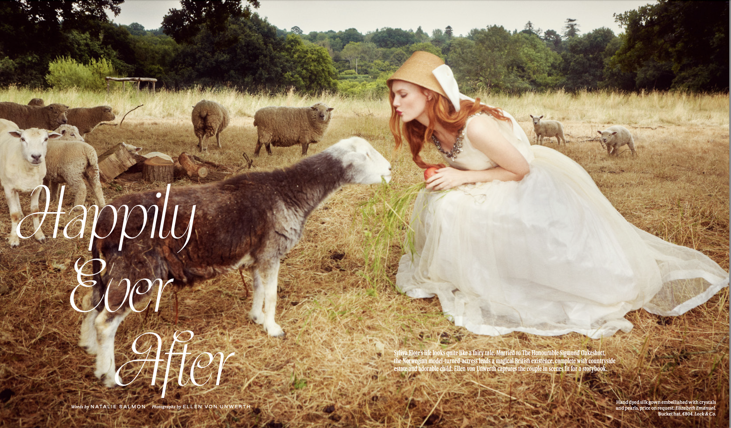 A woman dressed as a fairy in a white gown and a brown hat kneels in a field of dry grass, feeding a sheep. In the background, there are sheep and a lush green forest. The image has text overlay that reads 'Happily Ever After' and credits the photographer and designer.