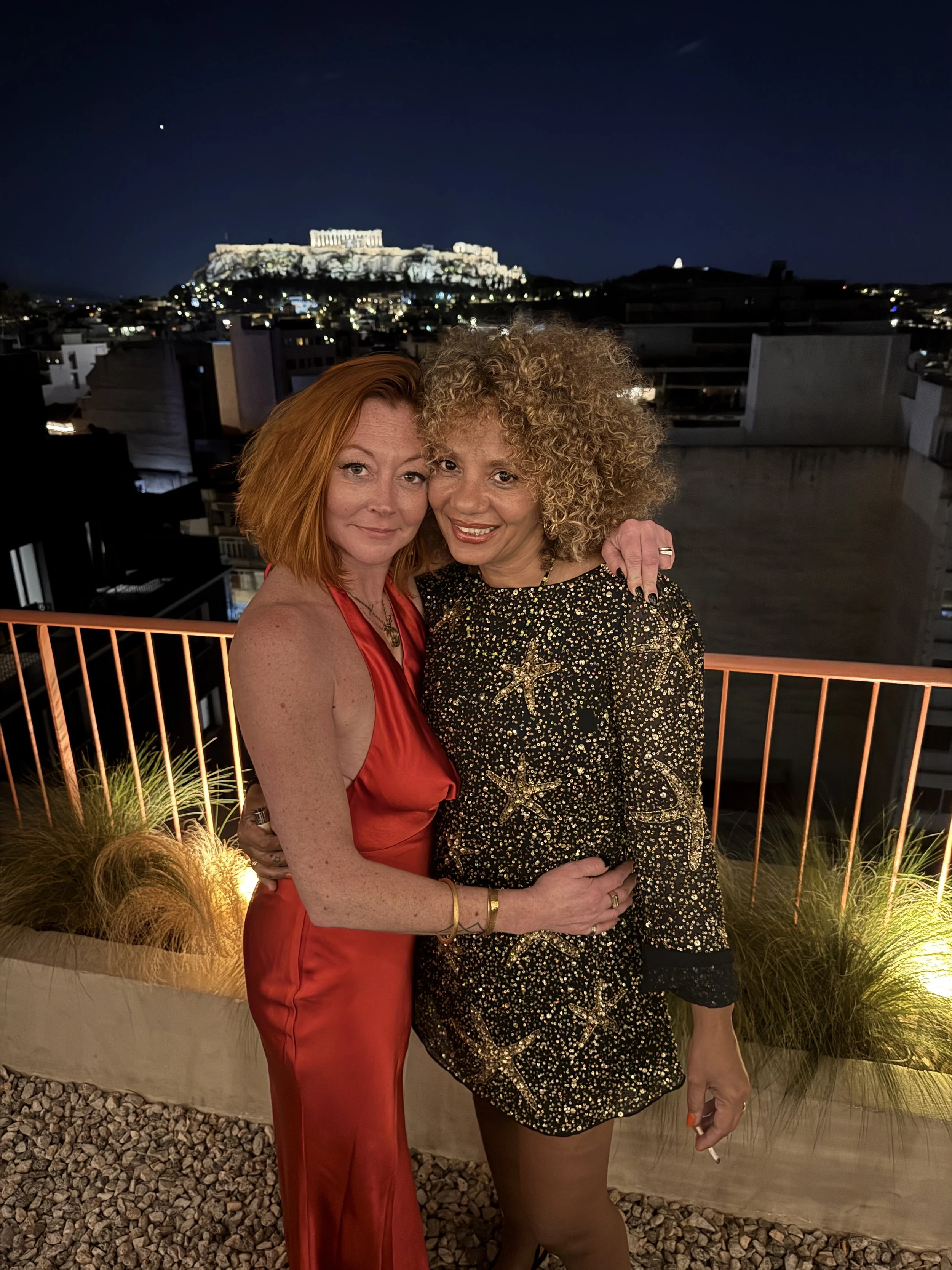 Two women posing closely together on a balcony at night with a view of the illuminated Acropolis in Athens, Greece, visible in the background.