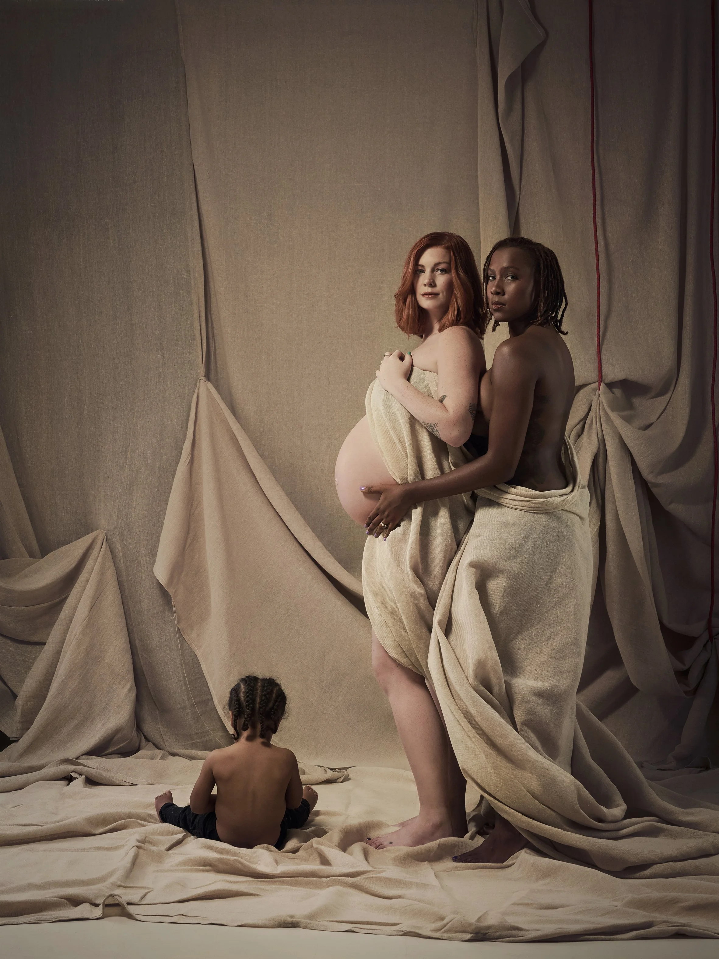 A pregnant woman with red hair standing in front of a beige fabric backdrop, holding her belly. A woman with dreadlocks and a child with braided hair sit on the floor in front of her, all partly wrapped in beige fabric.