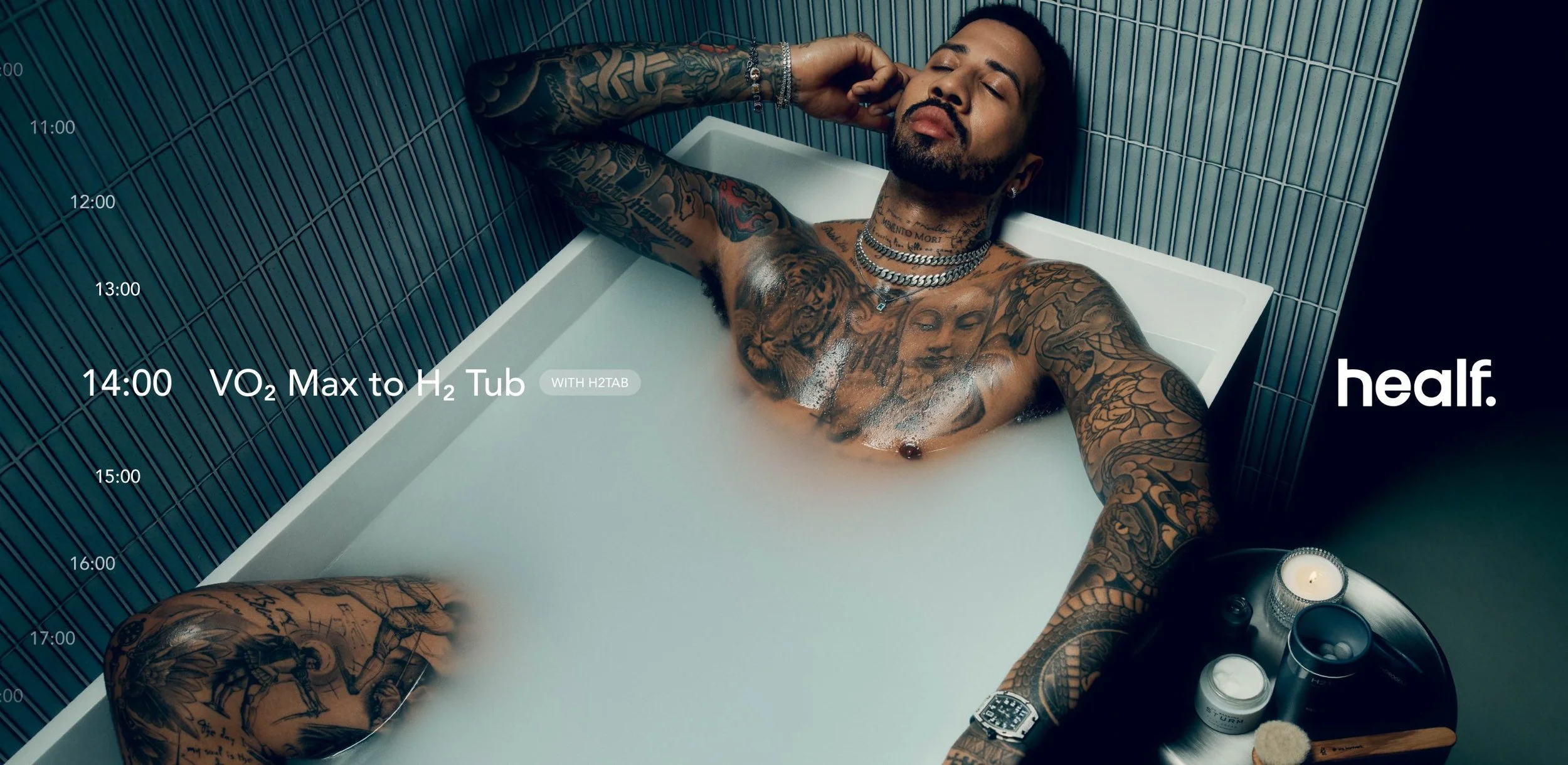 A tattooed man with closed eyes, lying in a bathtub filled with water and bubbles, surrounded by various skincare and beauty products, with a lit candle nearby.