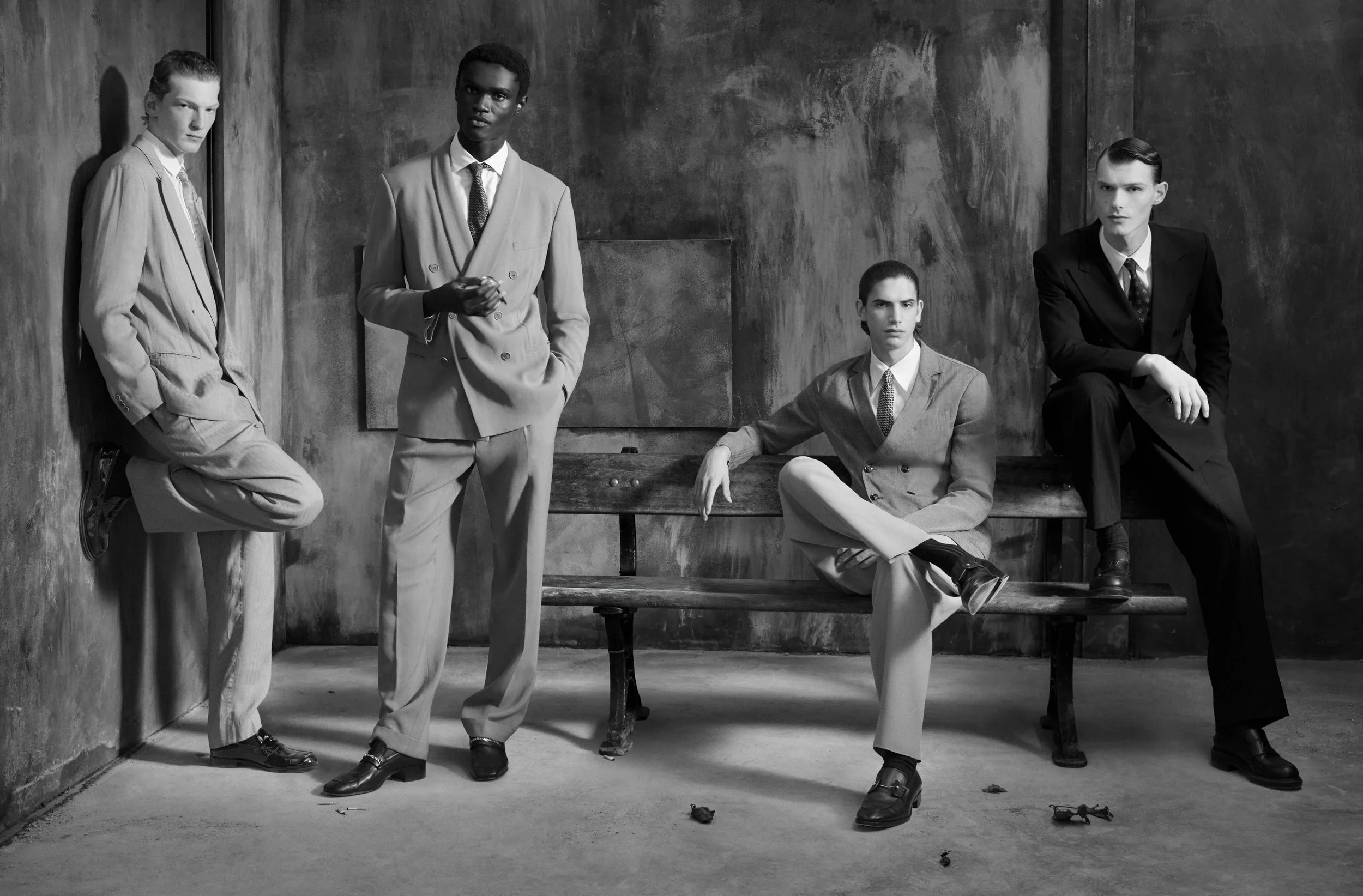 Black and white photo of five young men in formal attire posing against a concrete wall. Three are standing, and two are seated on a bench. The scene has a serious, stylish vibe.