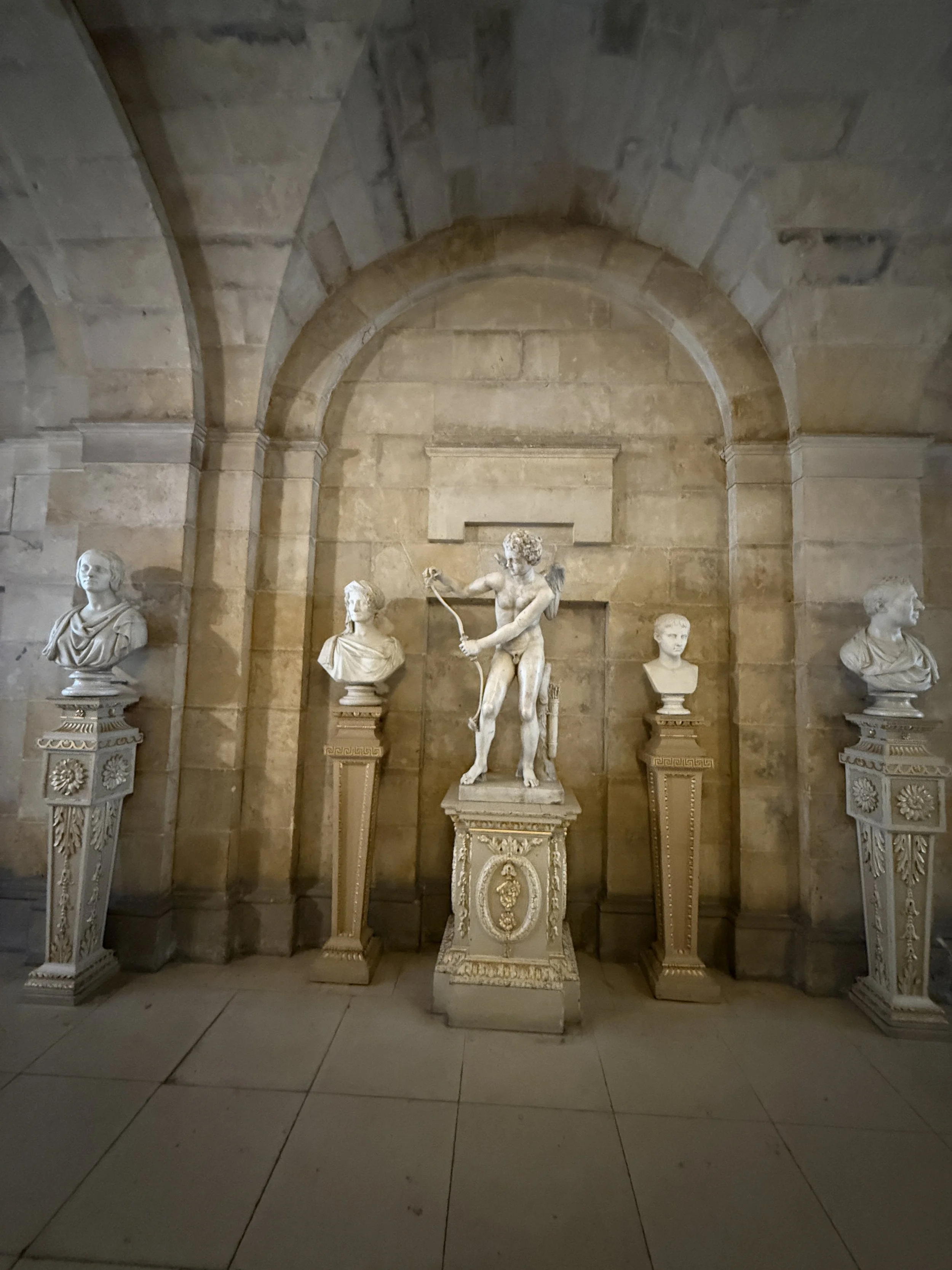 A marble statue of a young boy holding a bow and arrow, standing on an ornate pedestal in a stone-walled alcove, flanked by four bust sculptures on decorated pedestals.