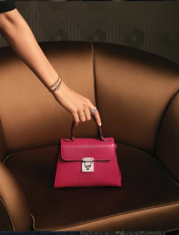 A person's hand with bracelets and rings points at a small pink handbag with a silver lock, placed on a curved brown leather sofa.