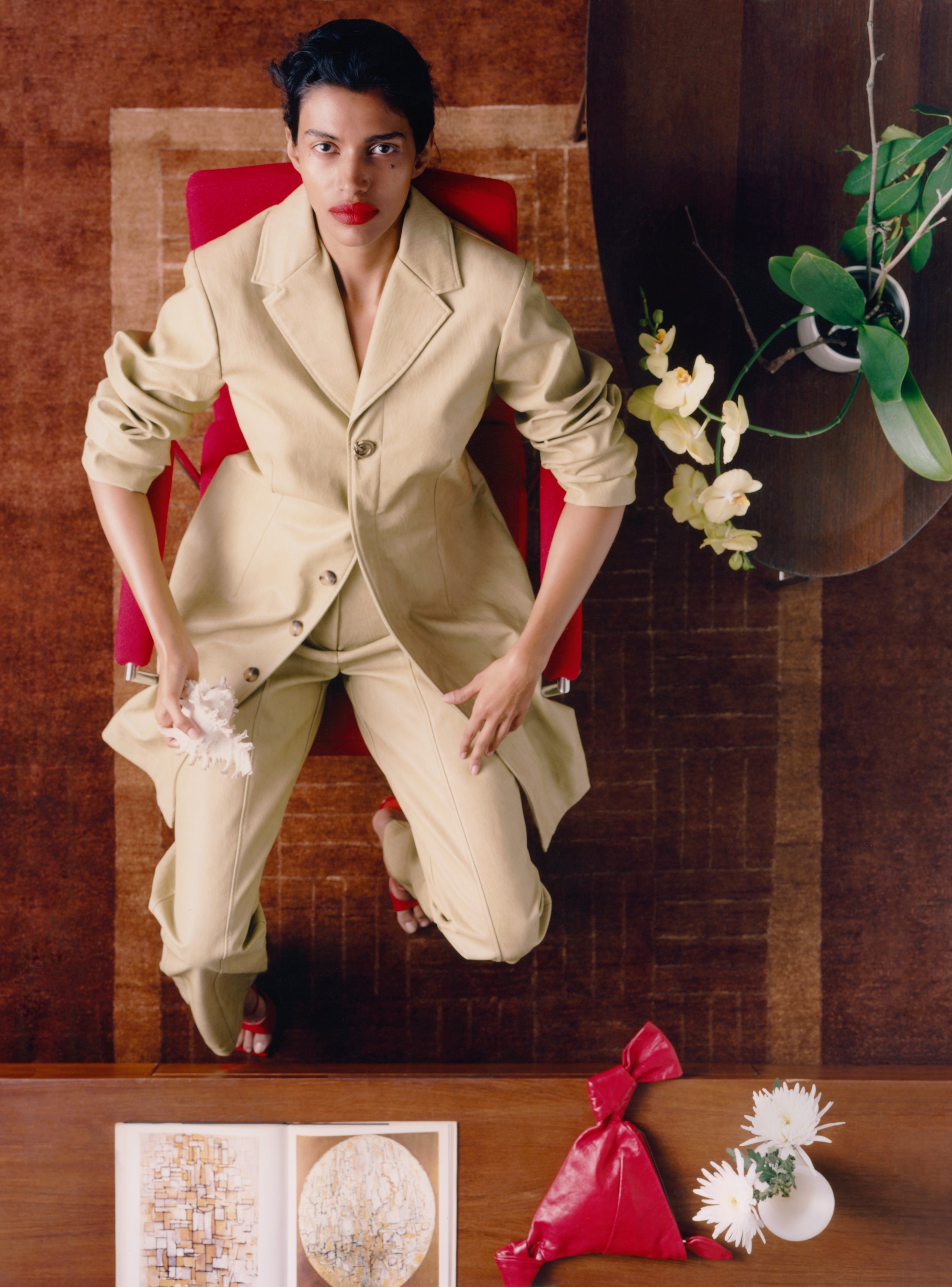 A woman with short black hair and red lipstick looking up at the camera, seated in a red chair at a wooden table, with a white vase containing flowers, a red wrapped gift, and art prints on the table.