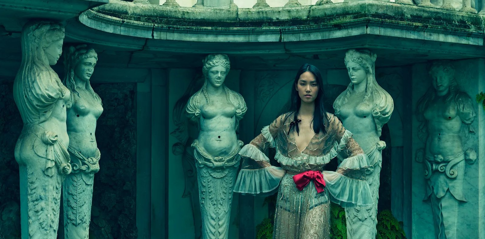 A woman in a detailed, sheer, gold dress with a red bow at the waist standing in front of a fountain adorned with classical sculptures of women.