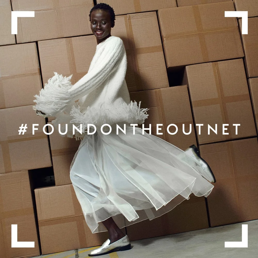 Person in a white sweater and flowing white skirt posing in front of stacked cardboard boxes, smiling.