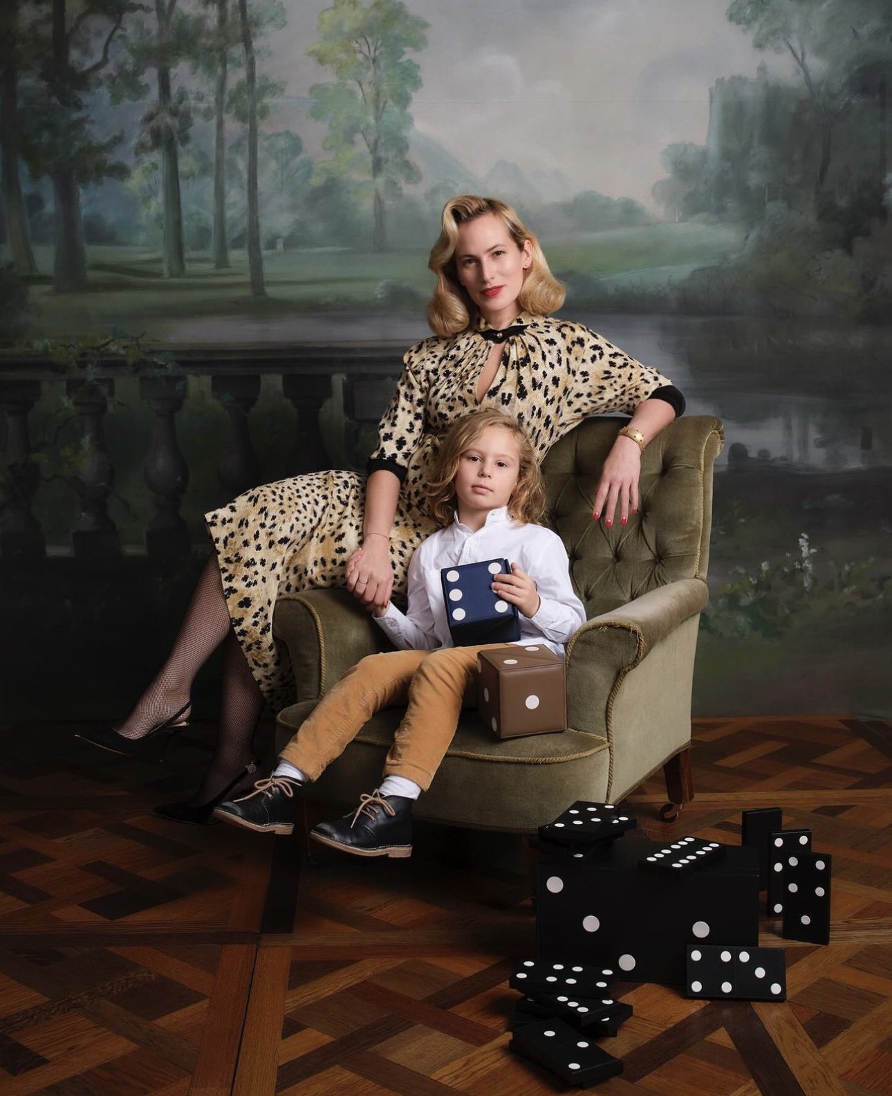 A woman and a young girl sitting on a vintage green armchair, with a painted outdoor landscape mural in the background. The woman wears a leopard print dress, and the girl wears a white shirt and tan pants, holding a large blue dice. Several big dominoes are arranged on the wooden floor around the chair.
