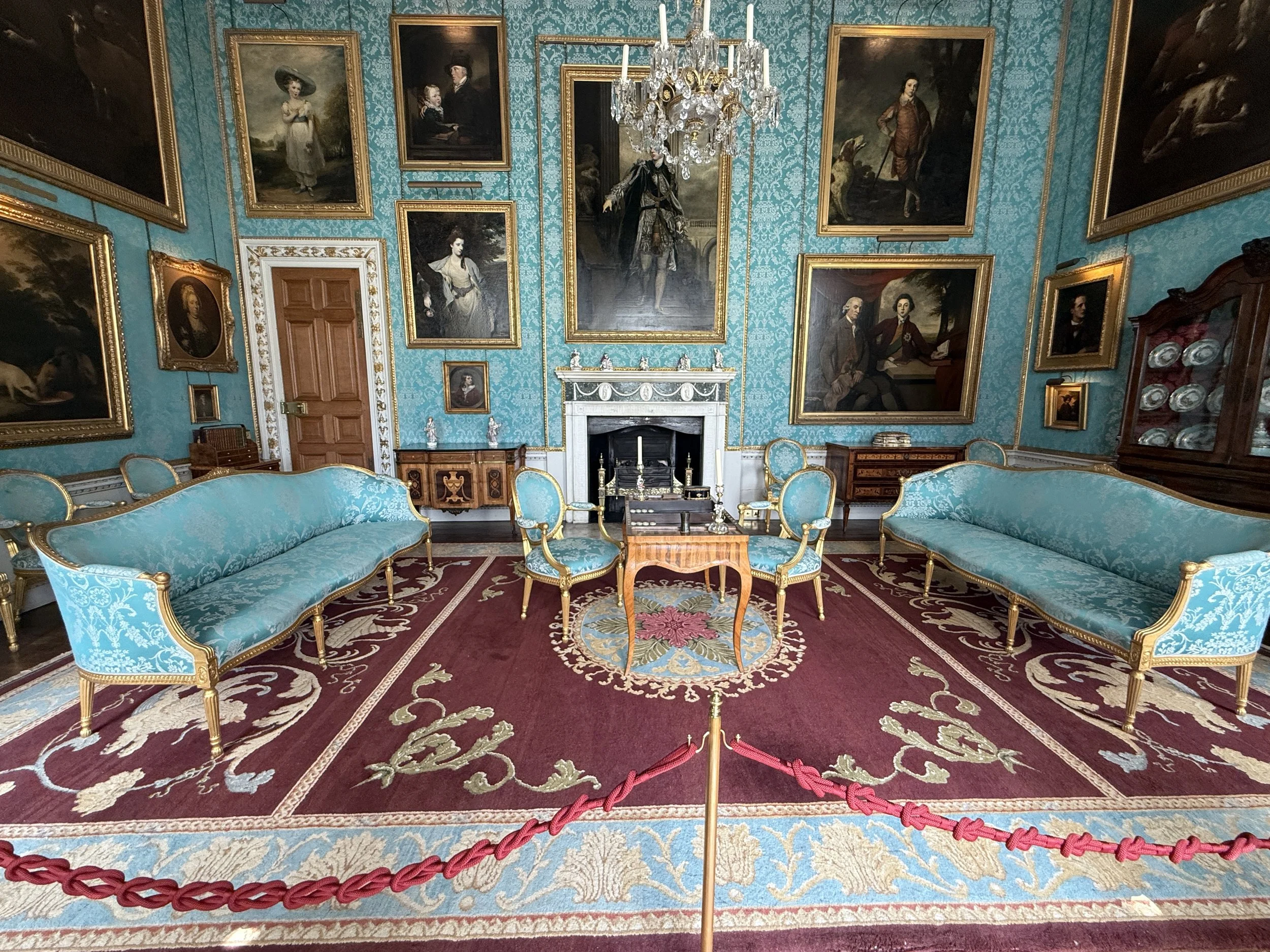 A room with teal patterned wallpaper and a red patterned carpet, furnished with light blue upholstered sofas and chairs with gold trim. The wall is decorated with numerous large, framed classical portrait paintings. There is a white fireplace with a black grate and a crystal chandelier hanging from the ceiling. A red velvet rope barrier is set up in front of the seating area.