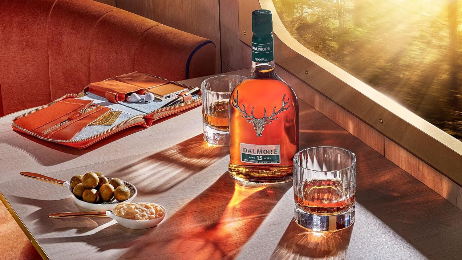 A table with a bottle of Dalmore 15-year-old scotch, two glasses with scotch, a bowl of green grapes, and a small bowl of potato chips. There are orange folders, a pair of white earphones, and a pen on the table. The background includes a window showing a landscape with water and trees, and part of a red velvet seat.