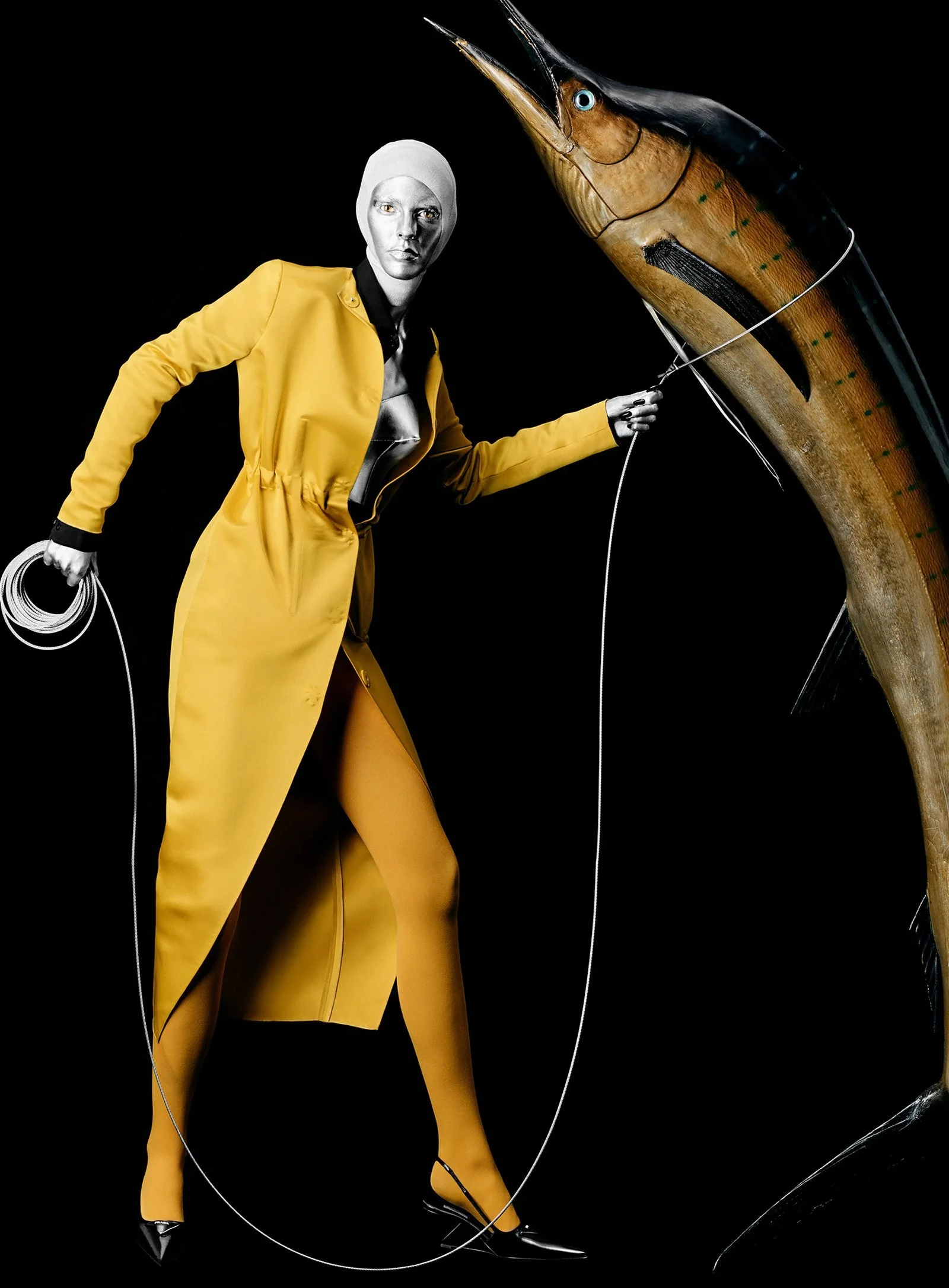 A person with a painted face wearing a yellow jacket and pants, holding a fishing rod, standing next to a large, mounted fish against a black background.