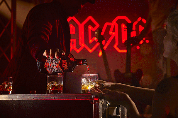 A bartender pours a drink from a bottle into a glass on a bar counter, with a woman reaching for her drink in front of a bright red neon sign that says 'AC/DC' in the background.