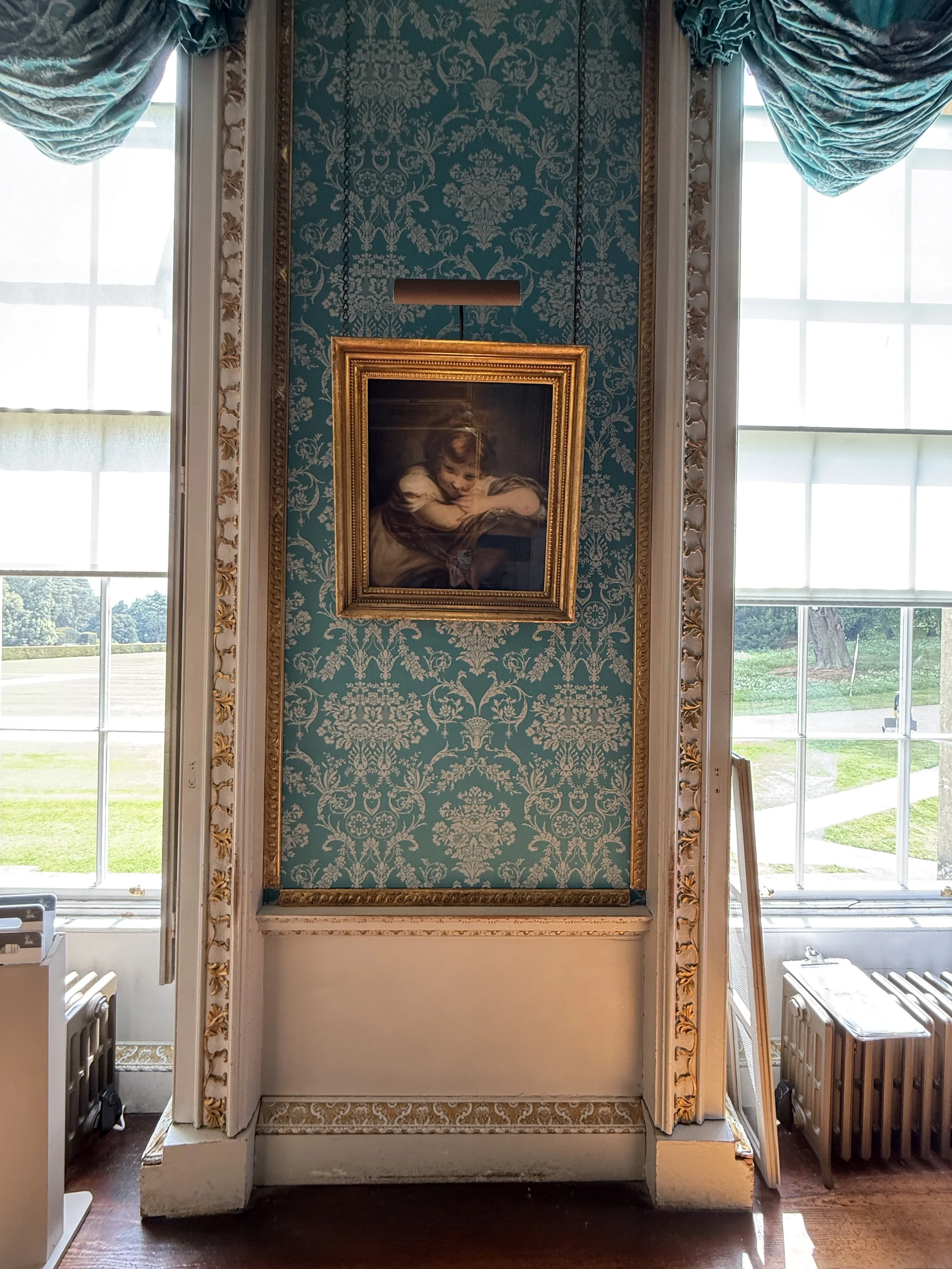 An ornate room with patterned teal wallpaper, gold-framed portrait of a young girl, large windows with blue draped curtains, and side tables with objects.