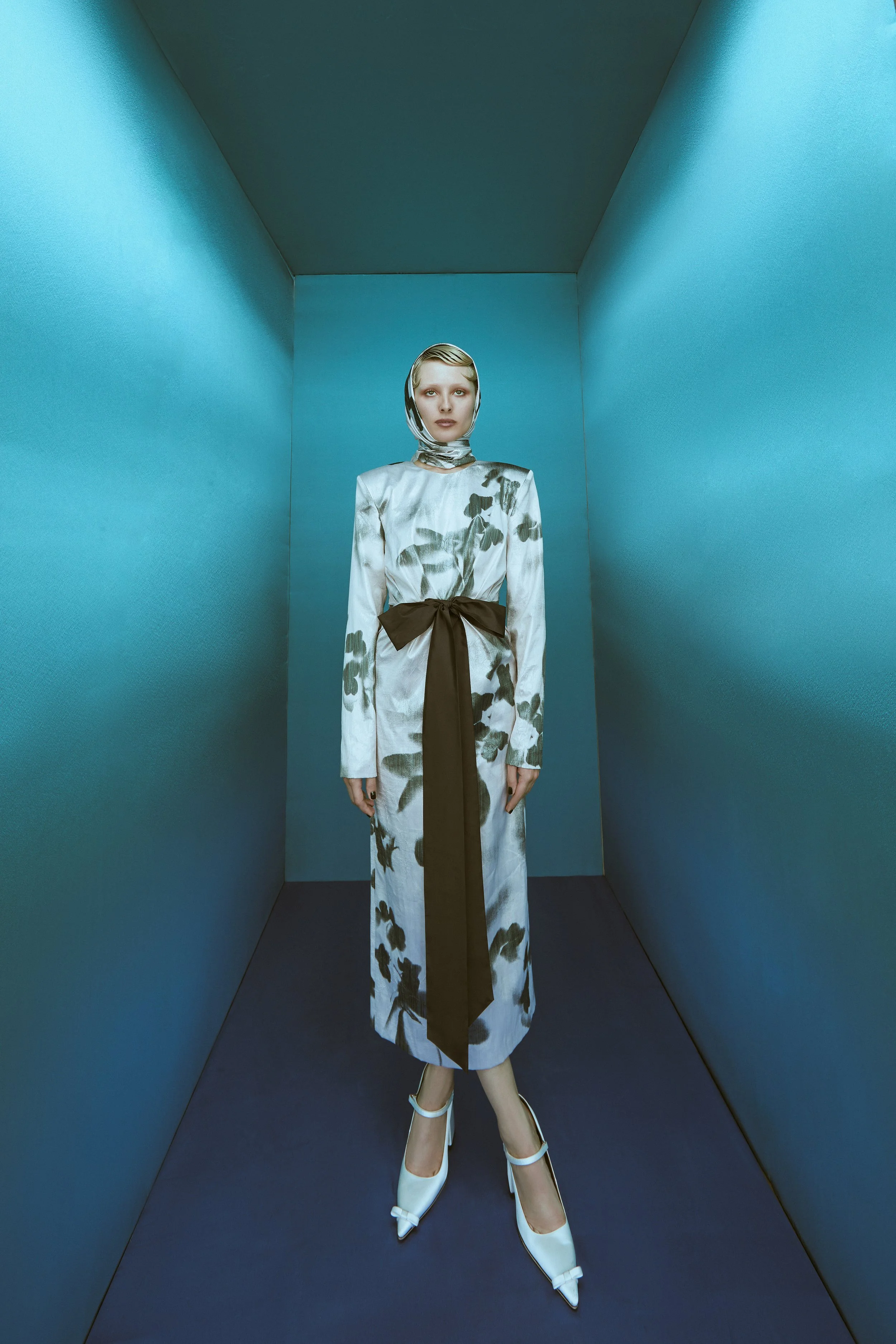 A woman stands in a narrow, blue-walled room, dressed in a floral patterned dress with a matching headscarf and white high heels.
