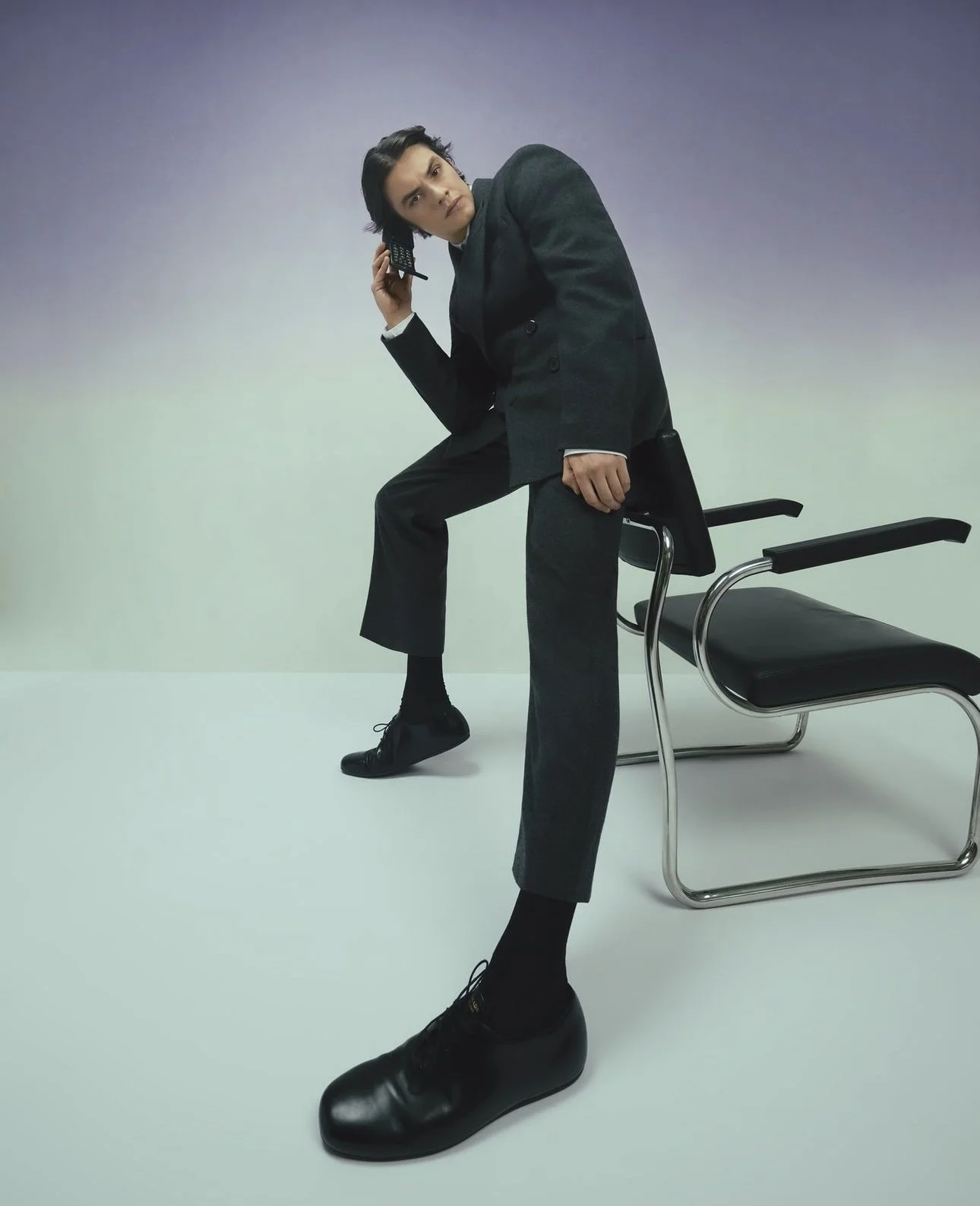 A man in a black suit is sitting on a black chair and holding a phone to his ear. His head is turned to the side and he has dark hair. The background is gradient from purple to light green.