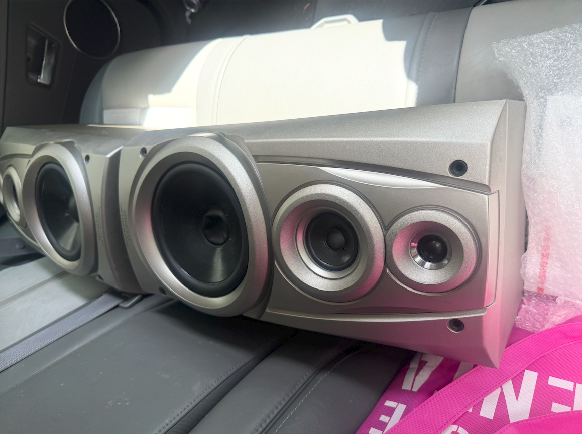 A large silver speaker with multiple drivers is placed on the back seat of a car, with some bubble wrap and a pink strap nearby.